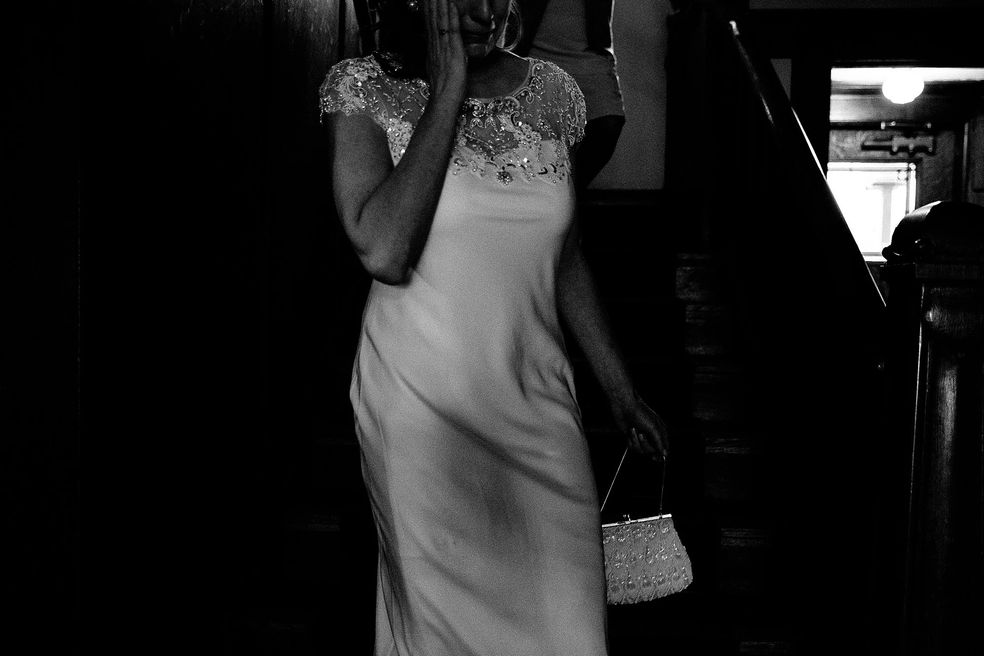 A woman in a light-colored dress with intricate embellishments on the shoulders, holding a beaded handbag, walking indoors near a staircase.