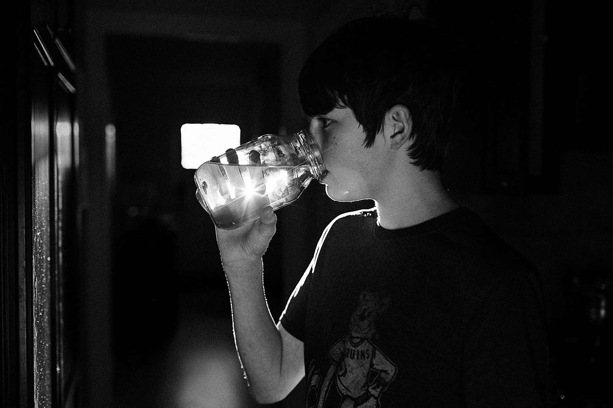 A person with short hair taking a sip from a plastic water bottle, standing indoors with a dark background and a bright window behind them.
