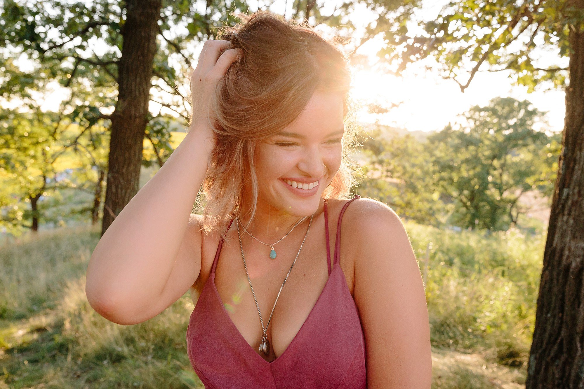 A young woman with reddish hair and a nose ring smiling outdoors in a forested area during sunset, wearing necklaces and a sleeveless maroon top.