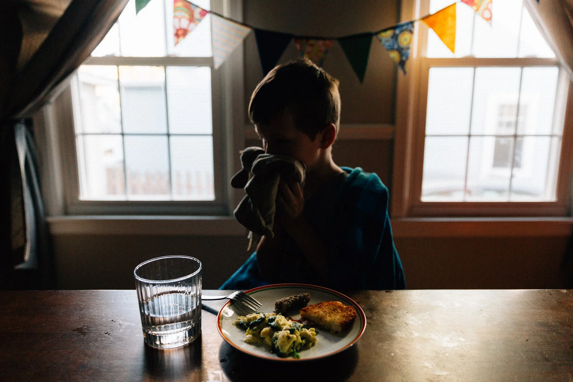 A young boy sitting at a dining table holding a cloth to his face, with a plate of food, including mashed potatoes and a piece of cooked meat, in front of him and a glass of water on the table. Behind him, colorful bunting decorations hang over a win