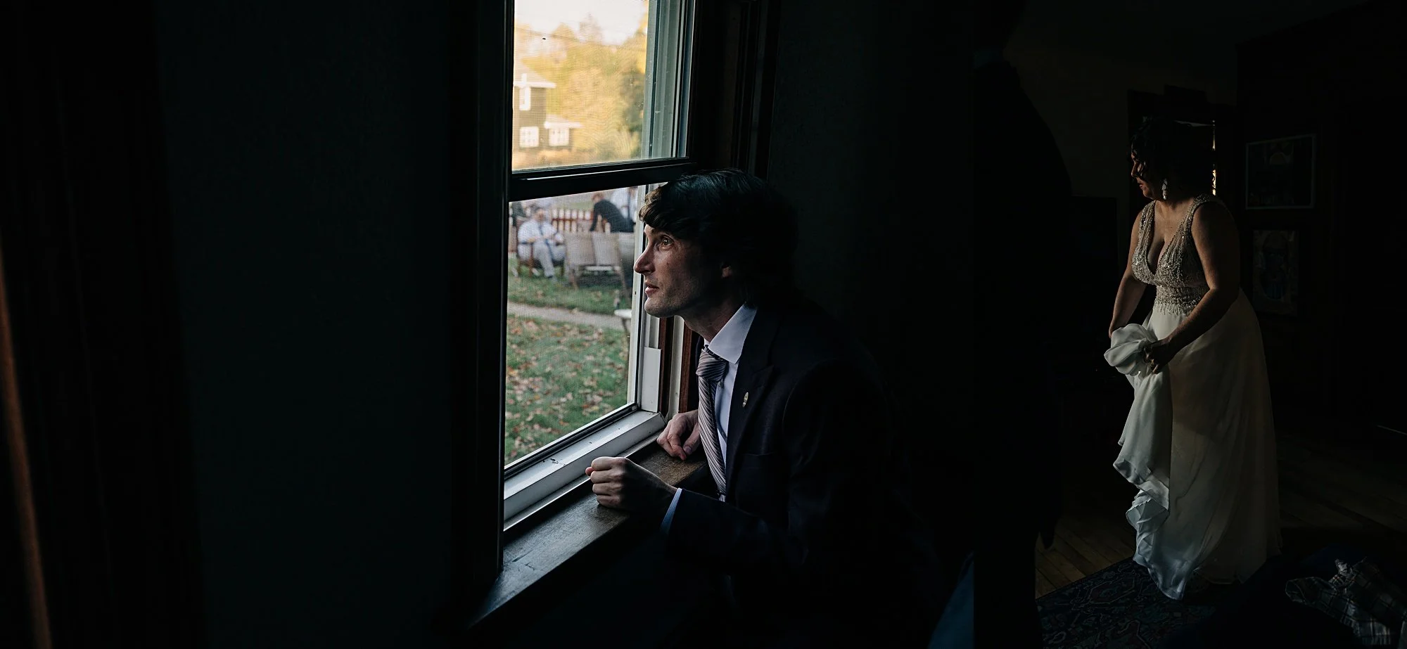 A man in a suit sitting by a window, looking outside with a thoughtful expression. A woman in a wedding dress is in the background, adjusting her dress inside a dimly lit room.