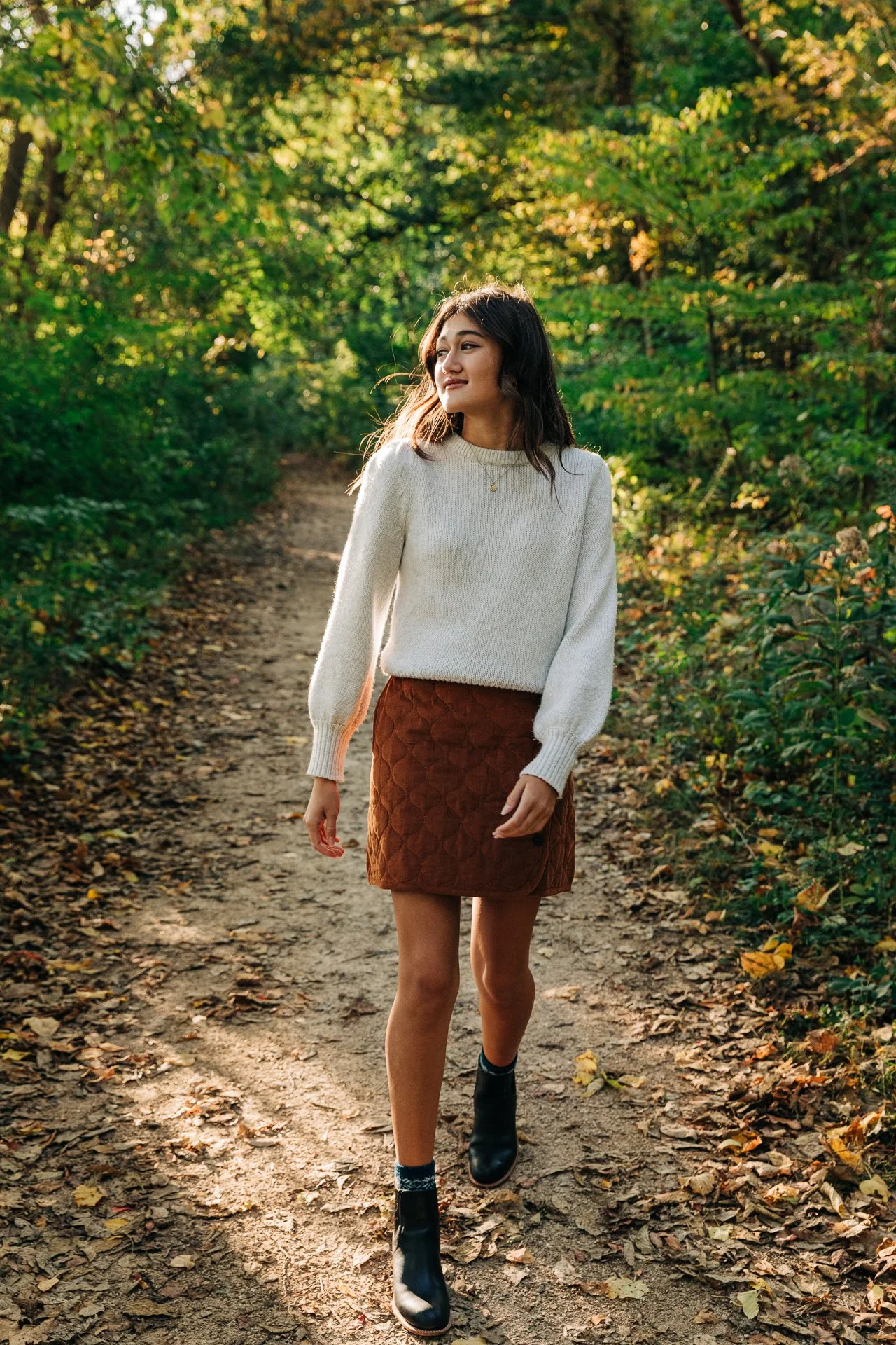 A young woman walking along a dirt trail in a forest, surrounded by green trees and foliage, during daytime with sunlight filtering through the leaves.