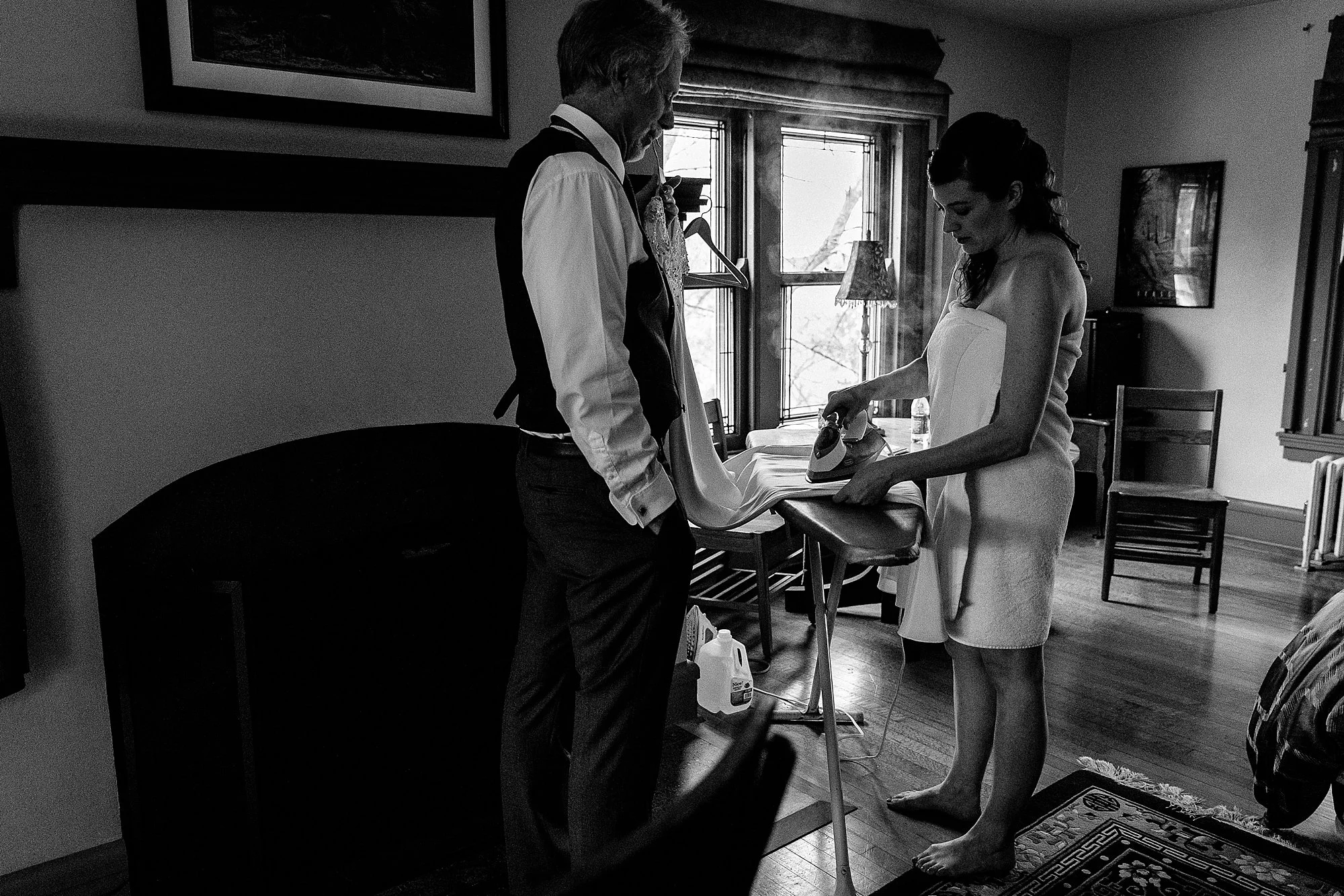 A woman in a towel is ironing a wedding dress in a room with wooden floors and vintage furniture, while an older man stands nearby, observing.