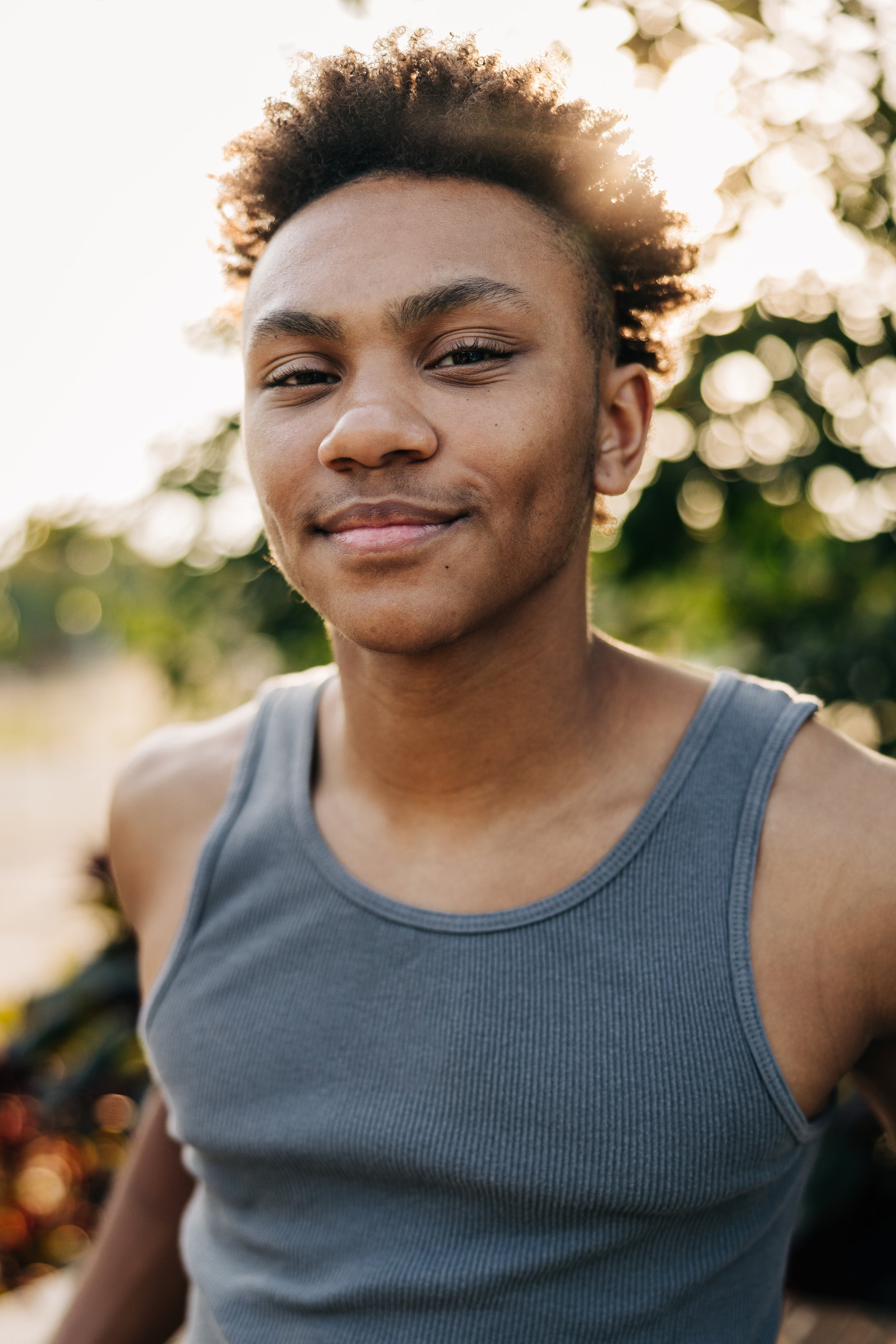 Young man with short curly hair wearing a gray tank top, smiling outdoors with sunlight filtering through trees.