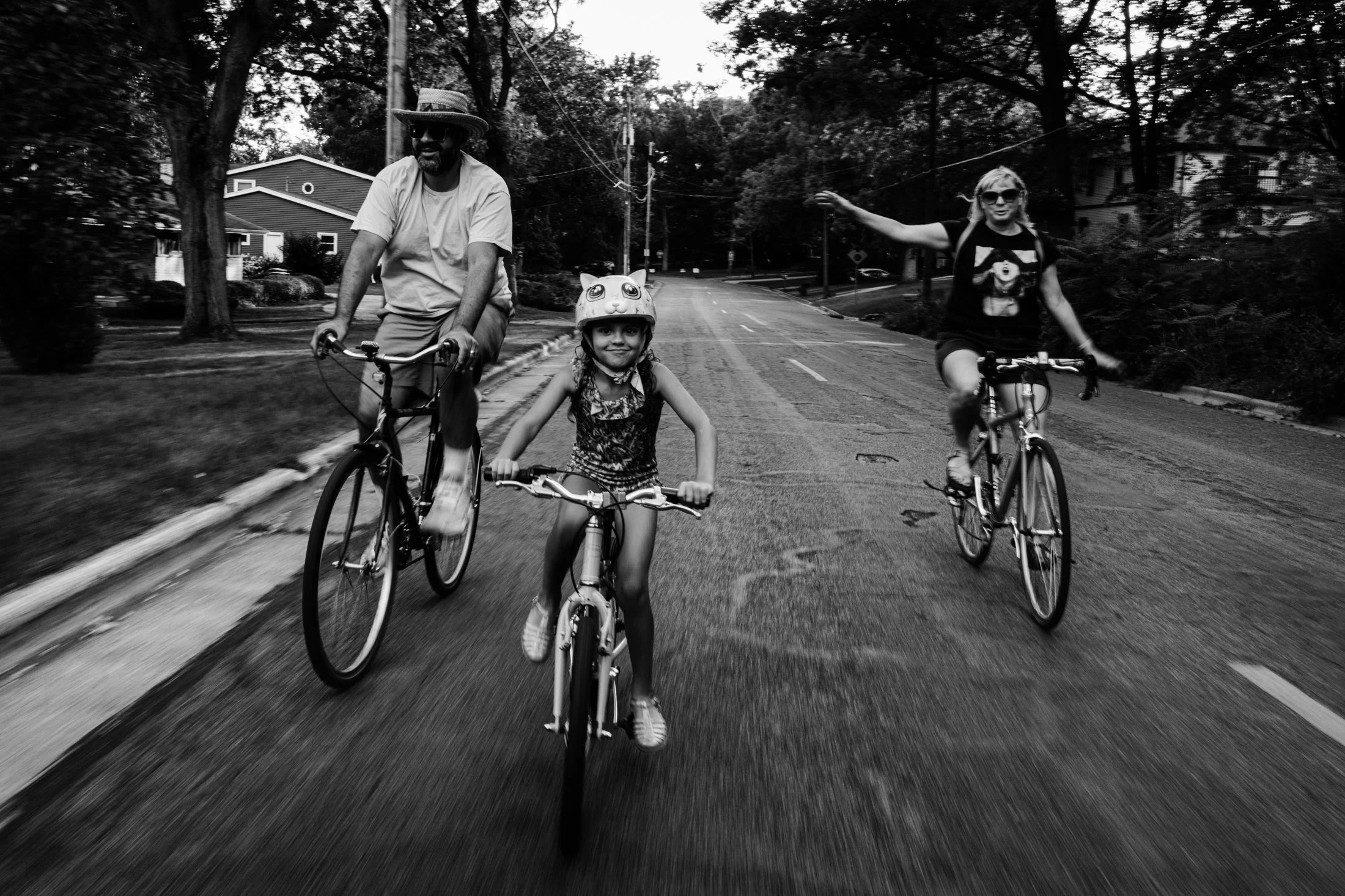 Three people riding bicycles on a suburban street, two adults and a young girl wearing a helmet with a cat face design. The girl is smiling, and the adults are cycling on either side, with one gesturing outward. The scene is set in a neighborhood wit