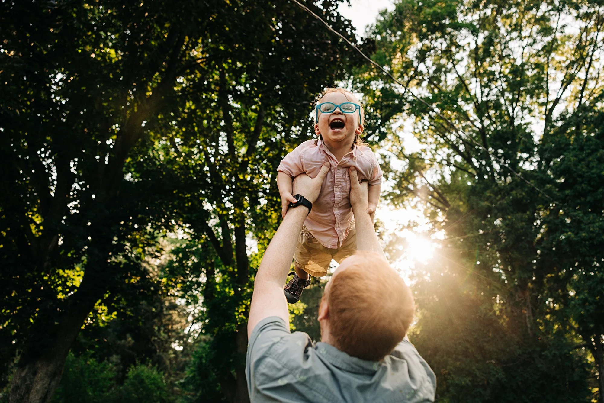 A man lifts a young girl into the air outdoors with trees and sunlight in the background, children wearing glasses smiling and laughing.
