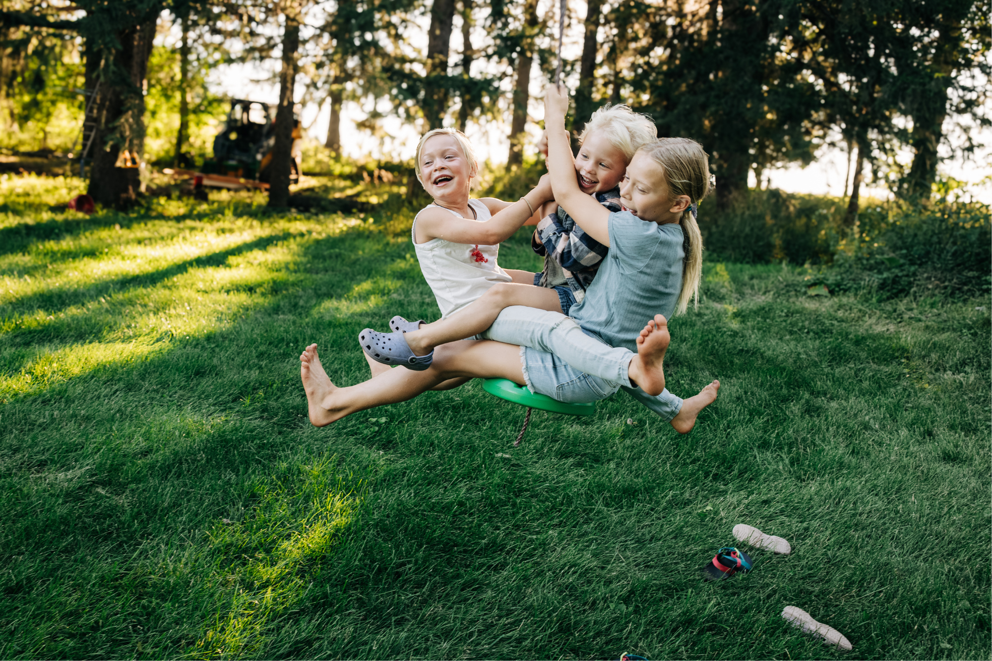 Three children playing on a green swing in a backyard with trees and sunlight.