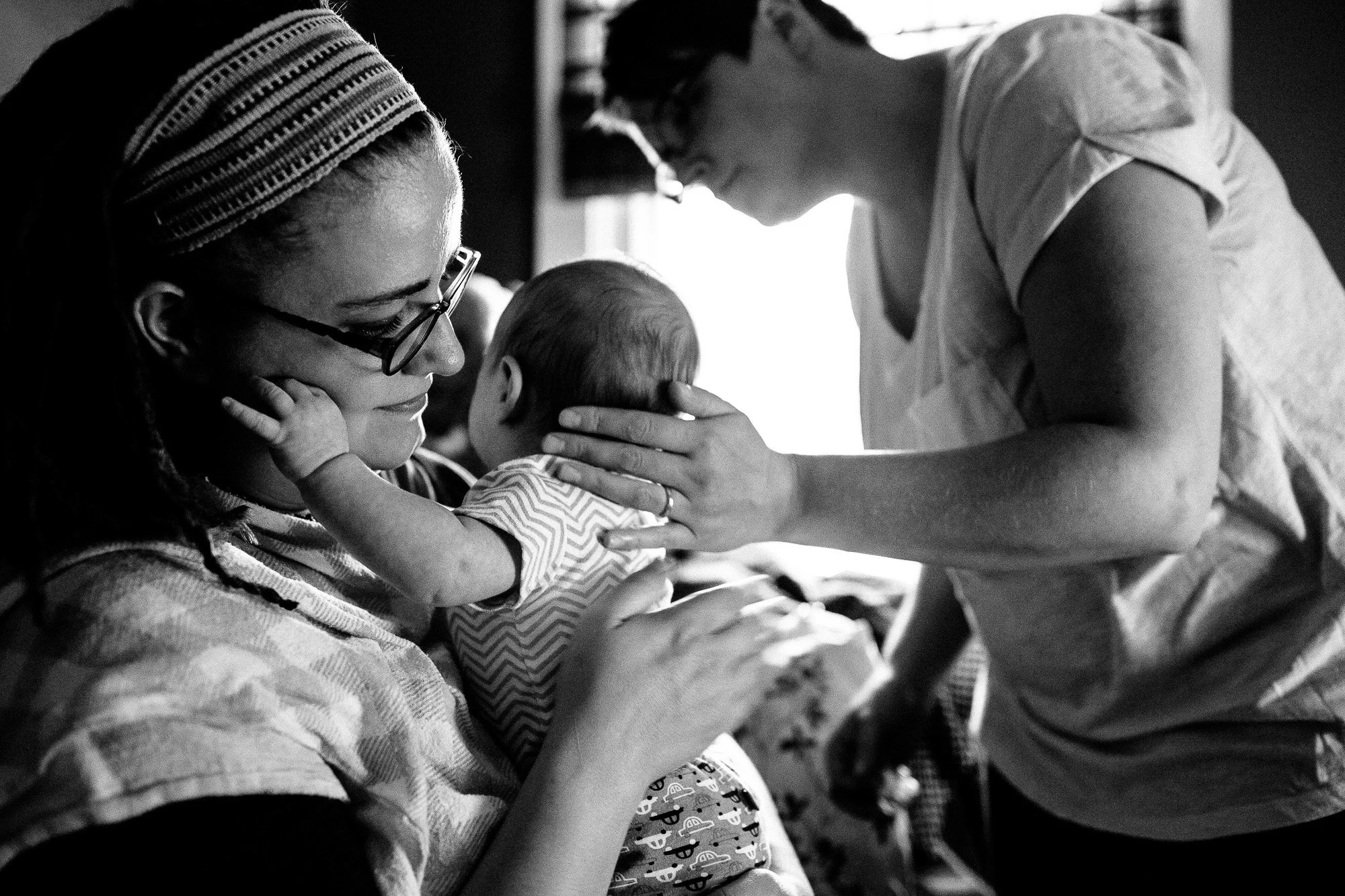 A woman with glasses and a headscarf holds a baby close, while another woman gently touches the baby's head, in a black and white setting.