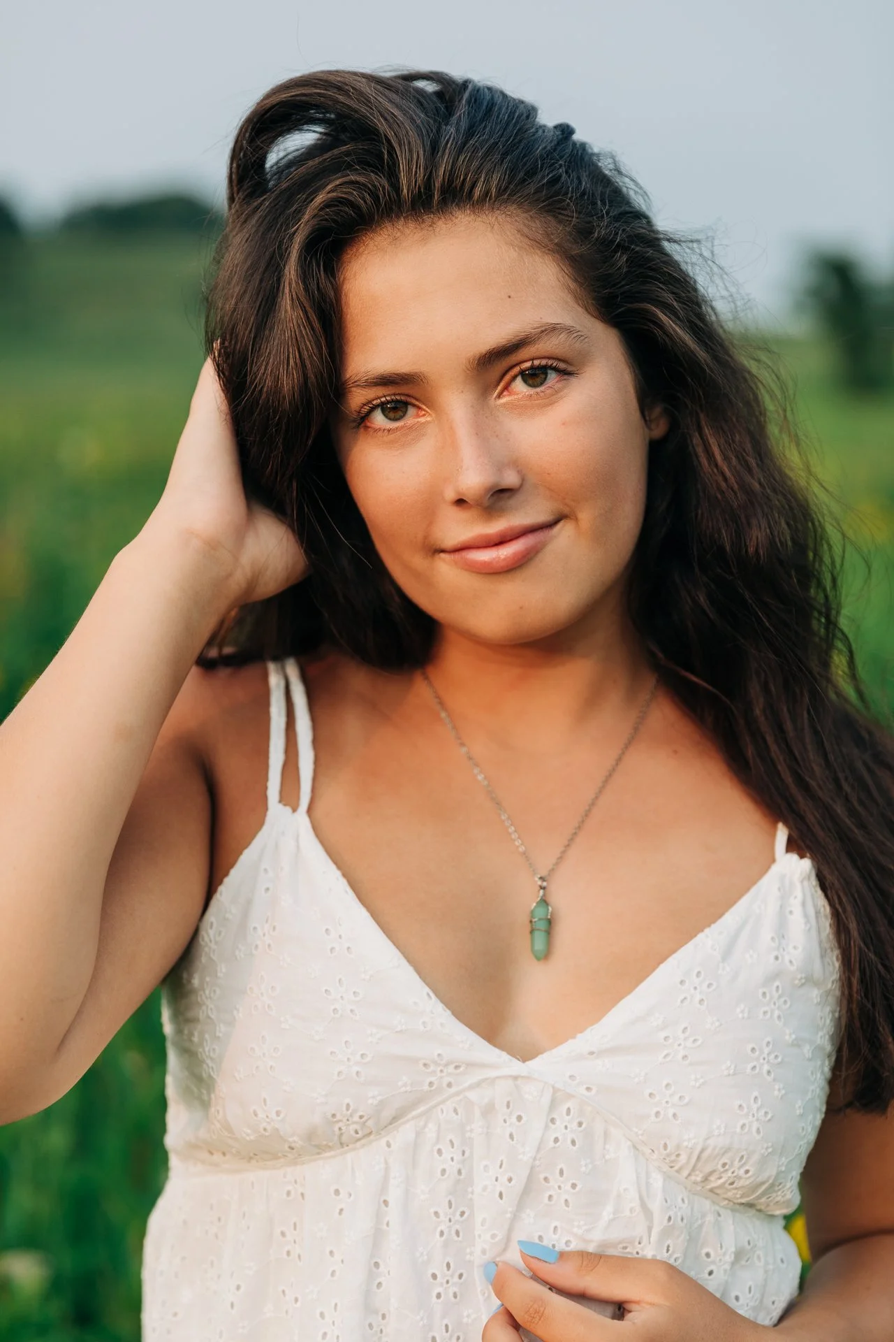A young woman with dark brown hair and light skin, wearing a white dress with eyelet details, a silver necklace with a green pendant, standing outdoors in a green field during daytime.