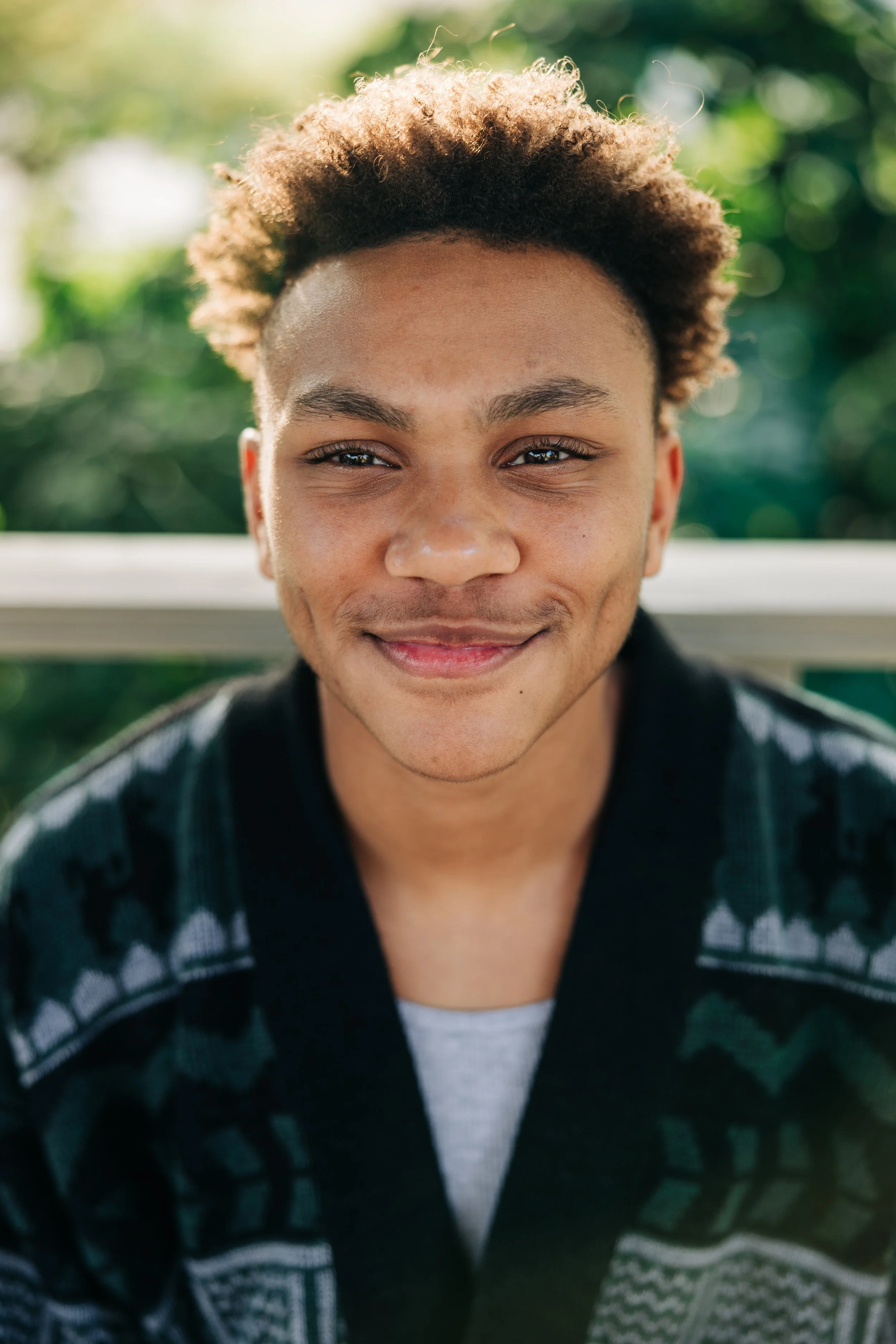 Portrait of a young man smiling outdoors with sunlight and greenery in the background.