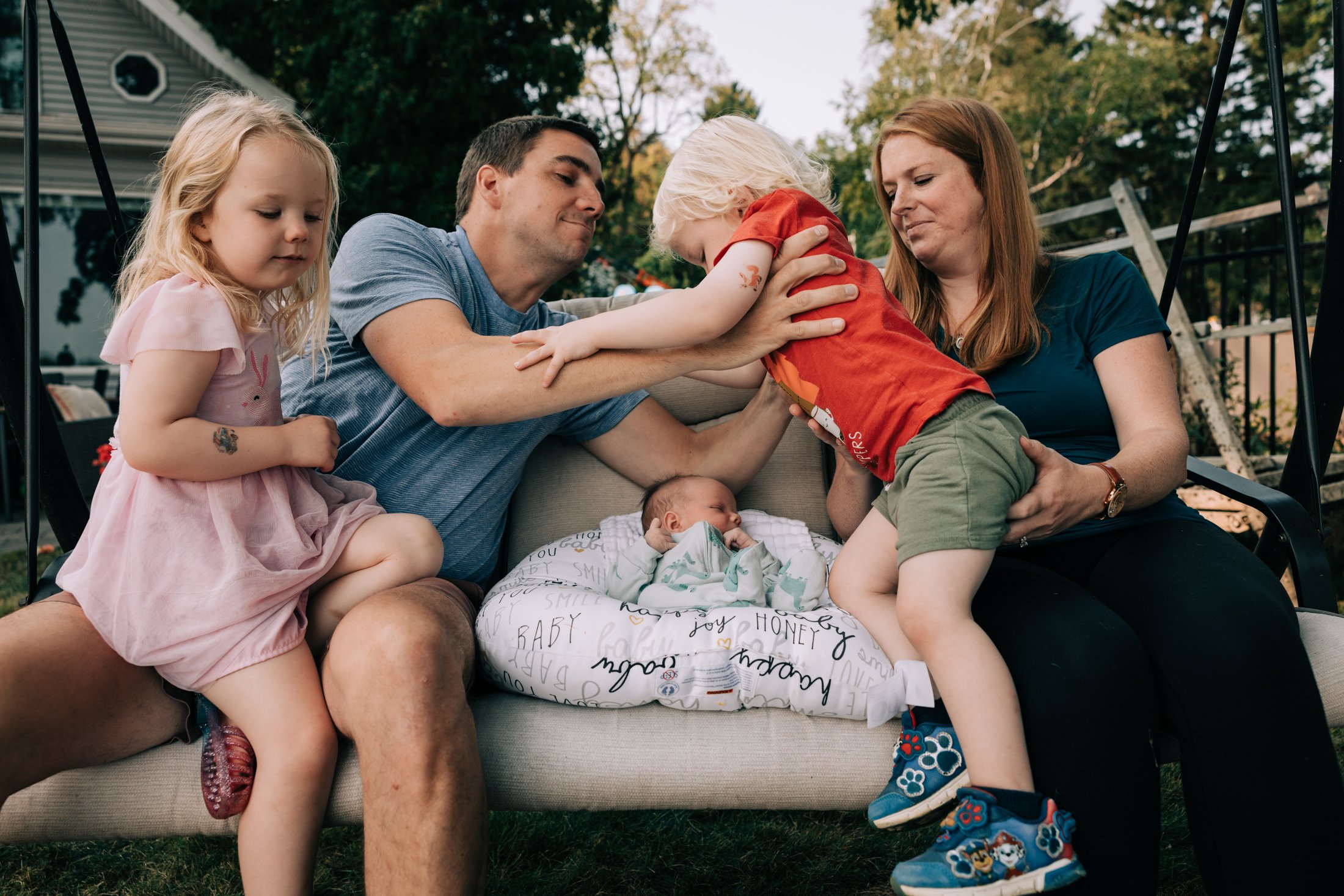 A family of five, including two young girls, a man, a woman, and a newborn baby, sitting on a outdoor couch in a backyard, playing and interacting with their children.