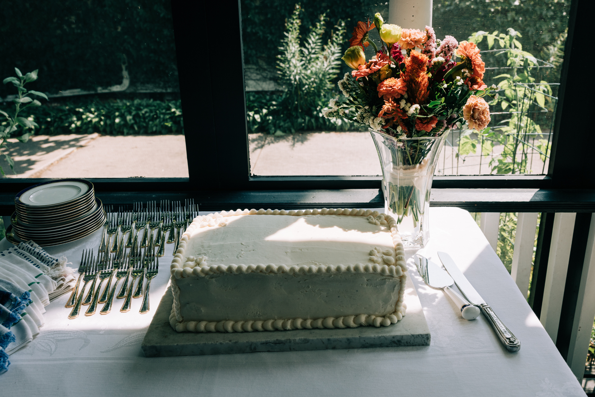 A rectangular cake with white frosting on a marble slab, decorated with piped borders, placed on a table with silverware, facing a window. There is a bouquet of pink, orange, and yellow flowers in a tall glass vase beside the cake. The table is set f
