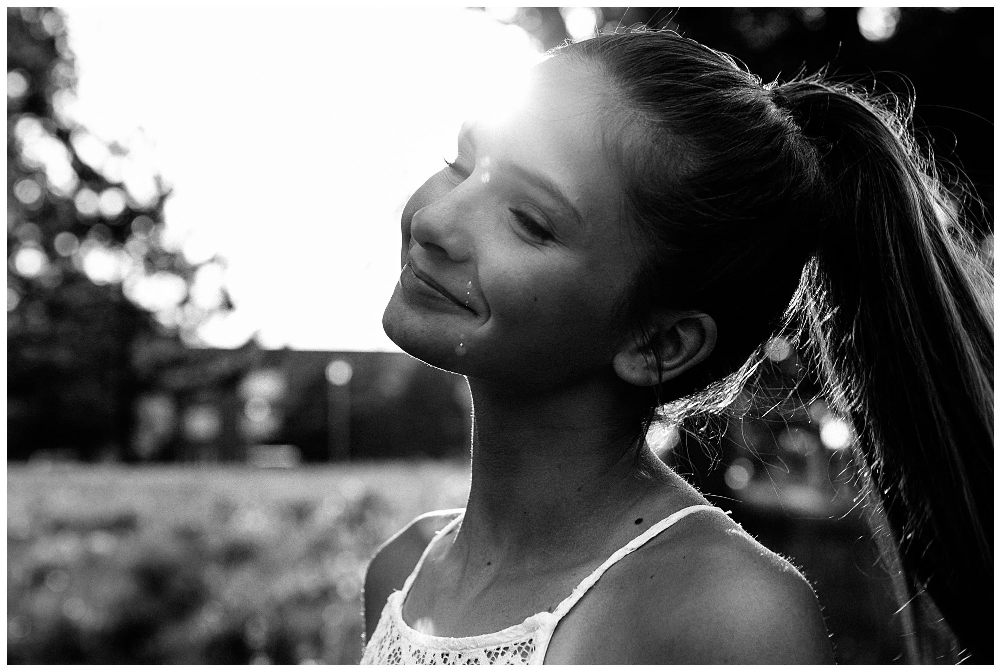 A young woman with a ponytail smiles with closed eyes outdoors during sunset