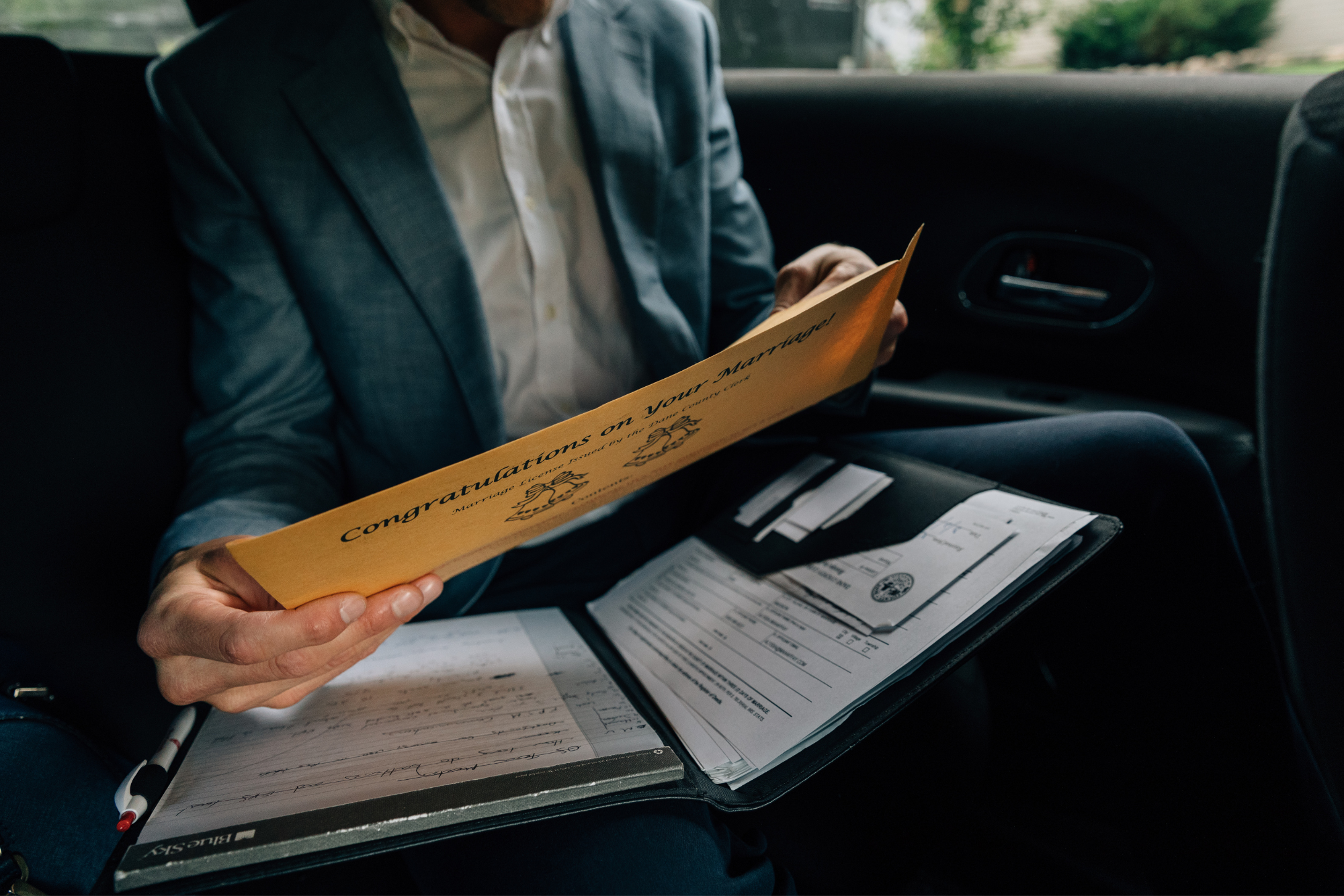 Person holding a yellow 'Congratulations on Your Marriage' card, sitting in a car with wedding documents in a folder on their lap.