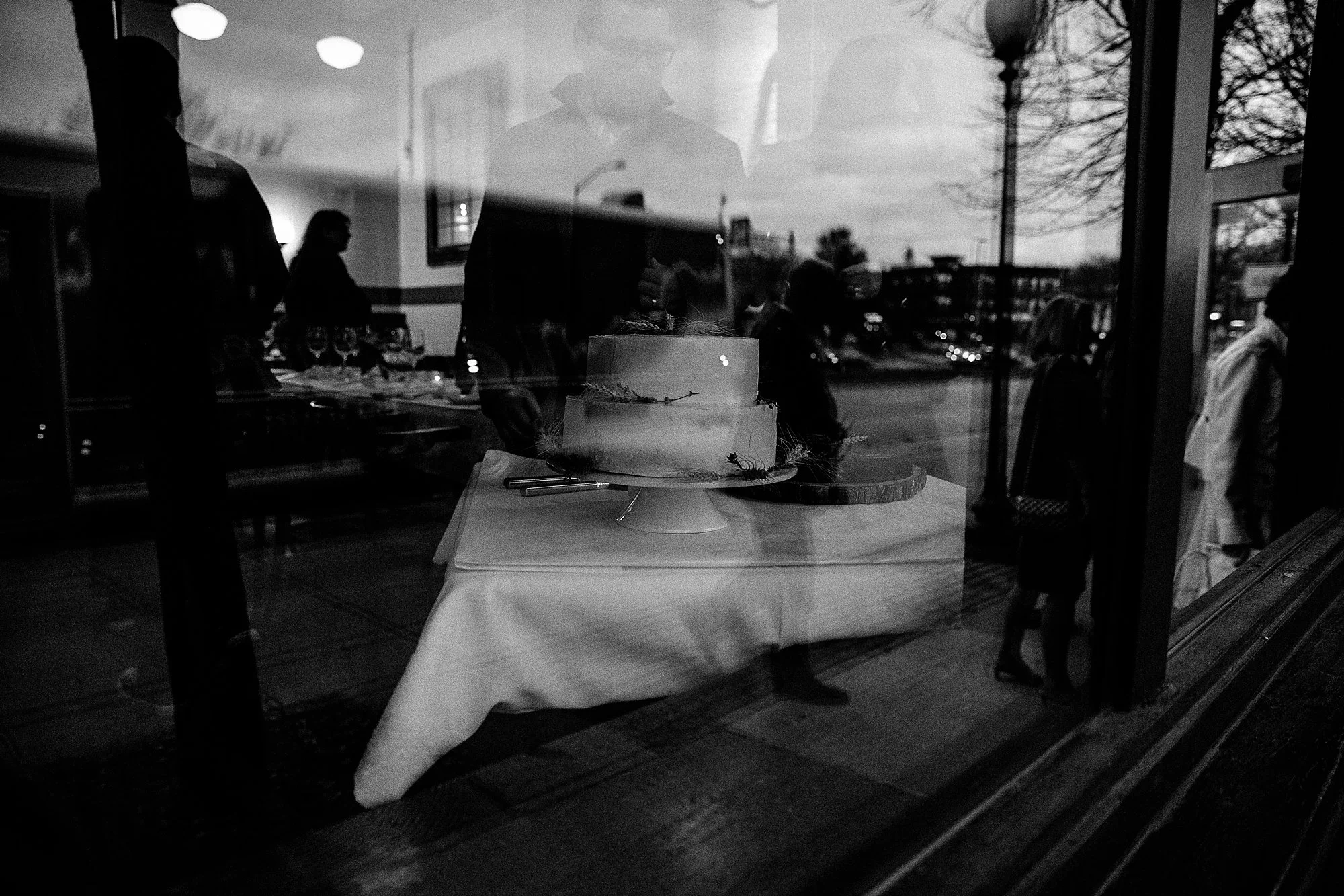 A cake displayed in a bakery window, with reflections of people and outdoor scenery visible on the glass.