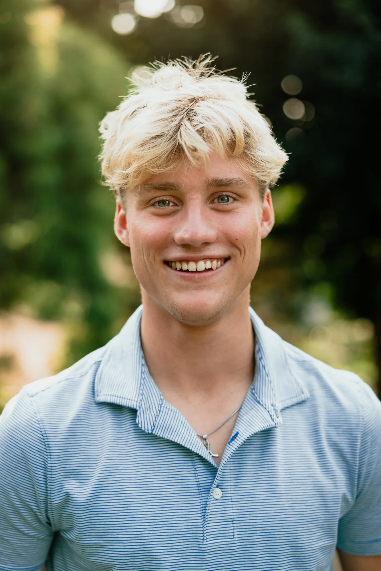 A young man with blond hair, blue eyes, and a bright smile, wearing a blue and white striped polo shirt and a silver necklace, outdoors with a blurred green background.
