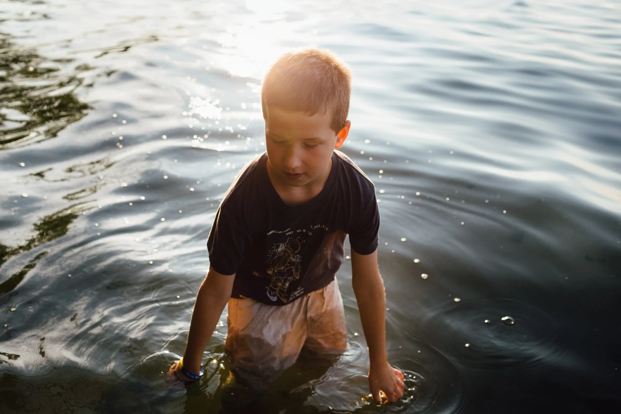 A young boy wading in a body of water during sunset, with sunlight reflecting off the water's surface.