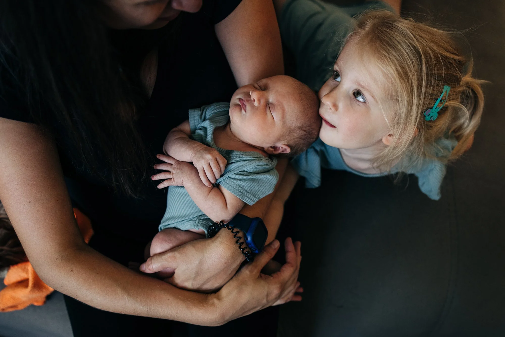 A young girl with blonde hair, wearing a blue clip in her hair, looks affectionately at a newborn baby in an adult's arms. The baby is sleeping, dressed in a striped outfit, and appears to be a few months old.