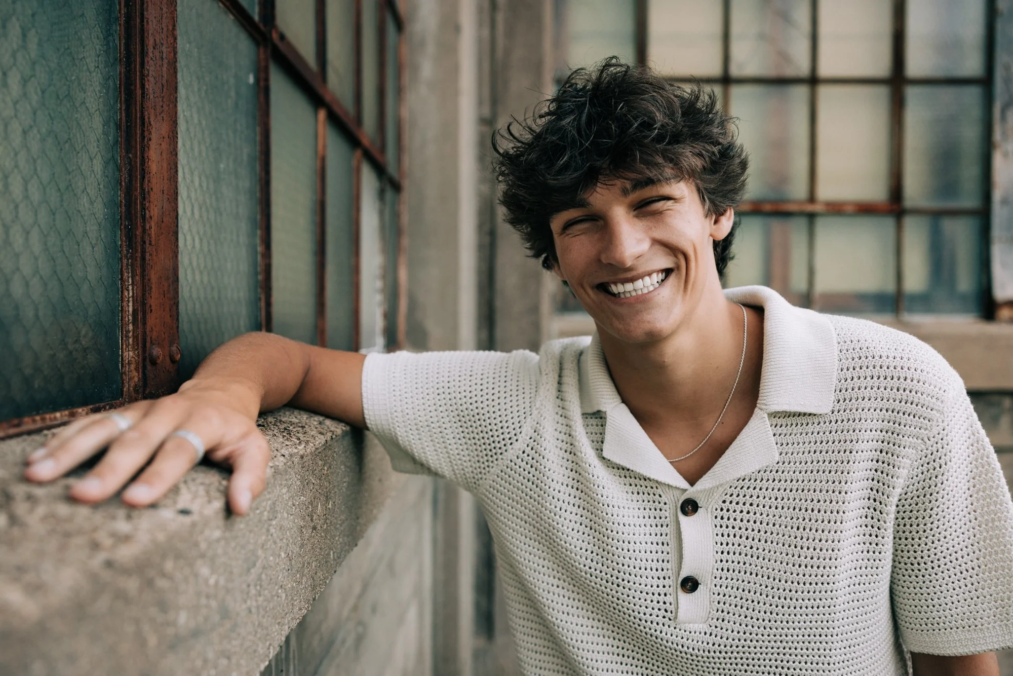 A young man with curly dark hair wearing a white knit polo shirt, smiling broadly, leaning on a concrete ledge, with industrial-style windows behind him.