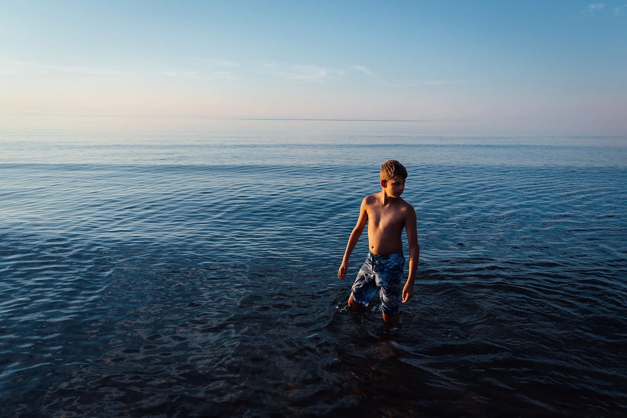 A young boy standing in shallow water at the beach during sunset, with calm ocean and sky in the background.