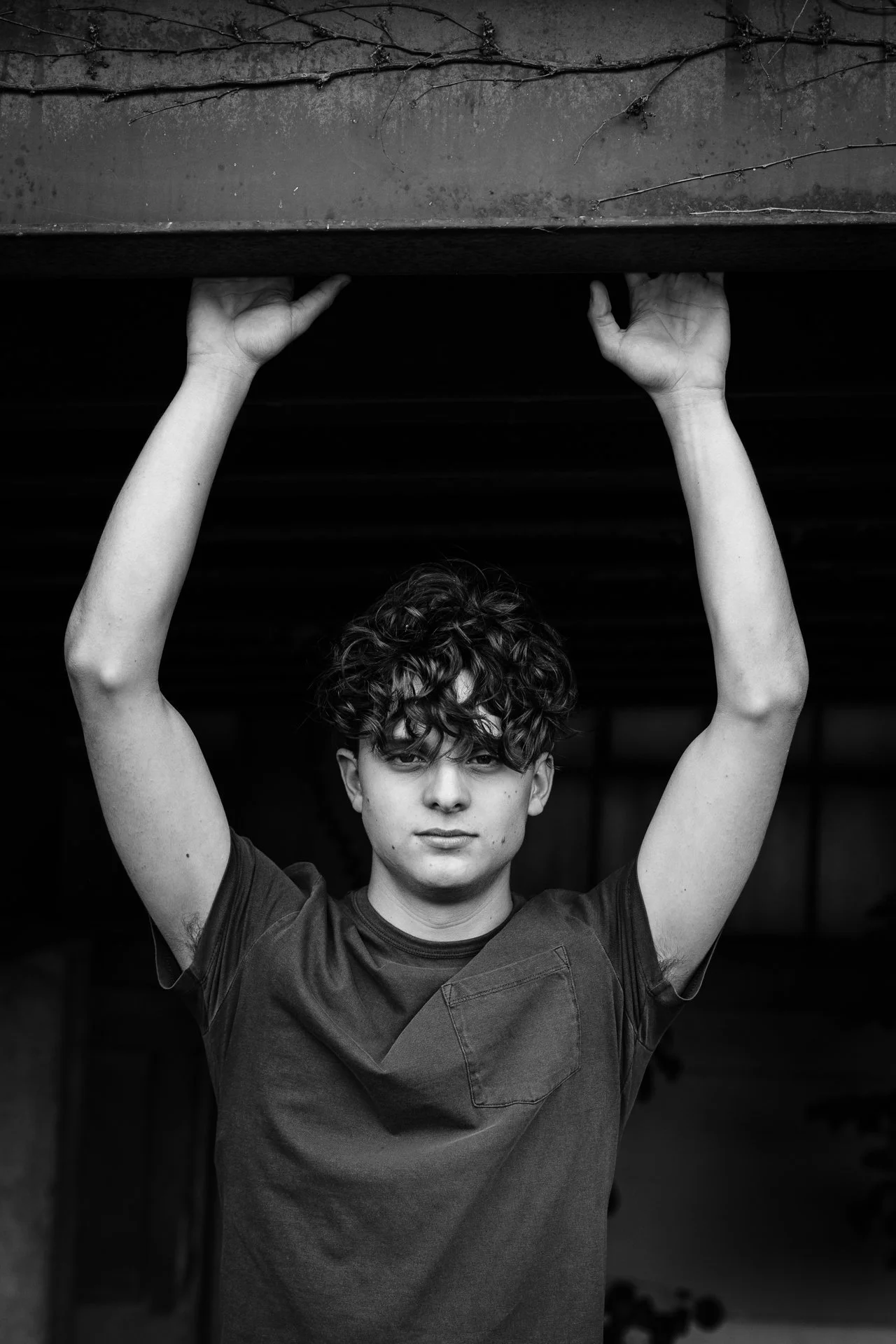 A black-and-white photo of a young person with curly hair, standing underneath a structure, holding onto the edge of the structure with both hands.