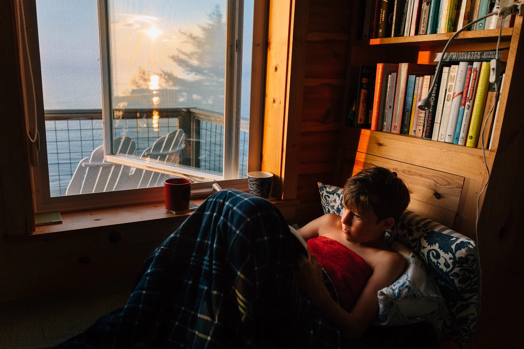 A boy lying in bed next to a window with a view of the sunset over a body of water. The boy is reading a book, with a soft pillow under his back and a red blanket. There's a bookshelf with various books, a lamp, and cups on the window sill.