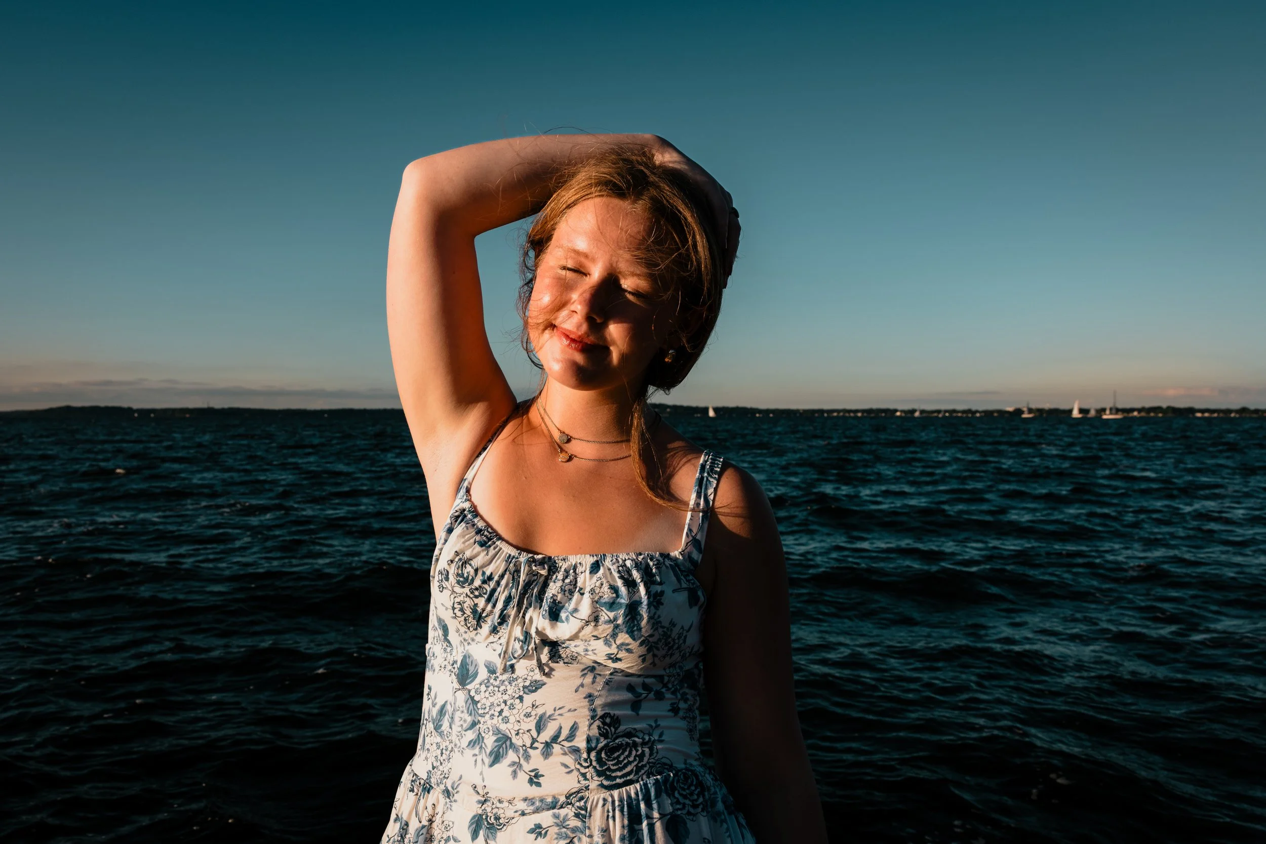 Young woman with closed eyes smiling, wearing a floral dress, standing by the water during sunset with sailboats in the distance.