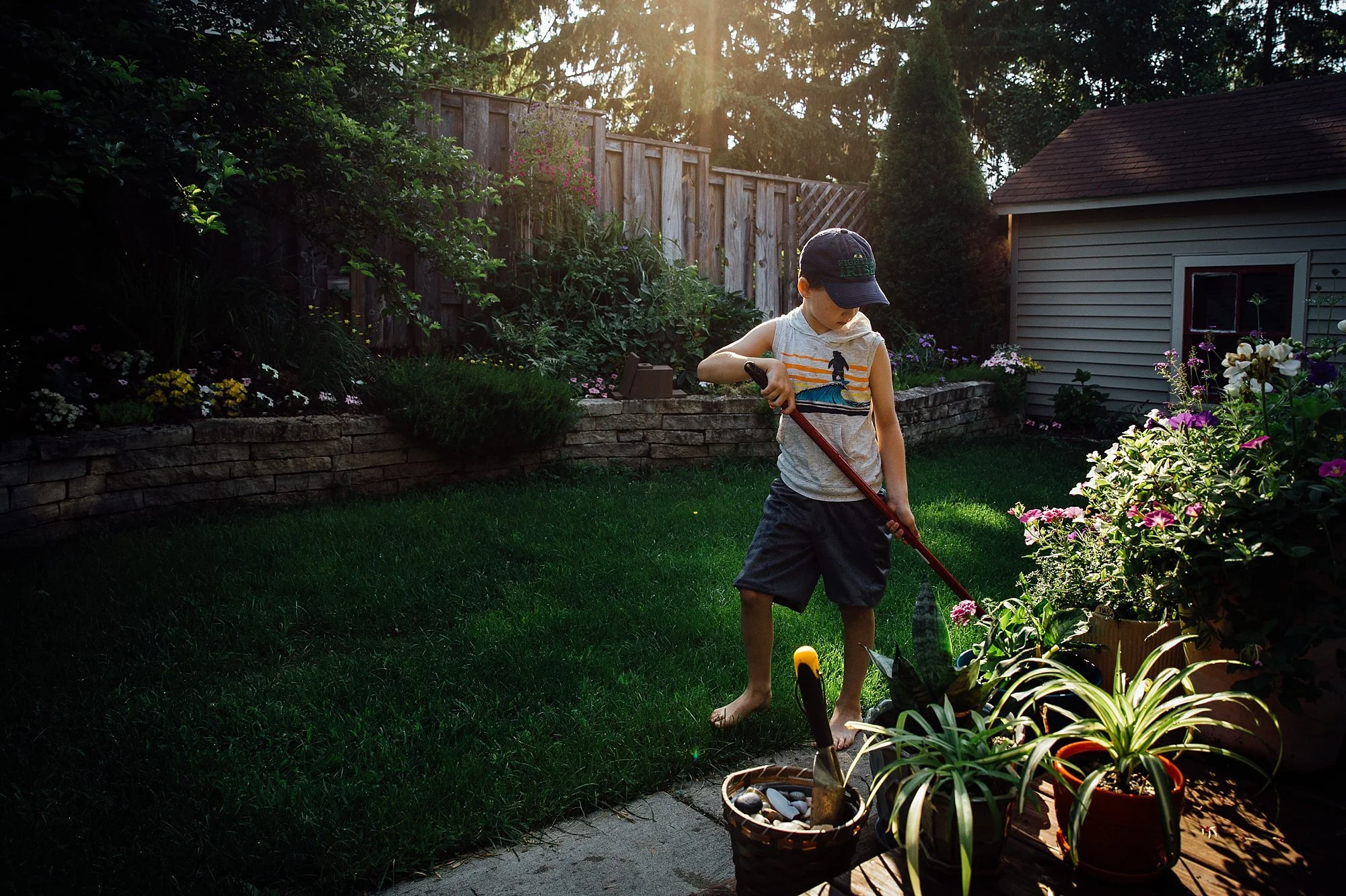 A young boy wearing a cap and sleeveless shirt is watering plants in a garden during late afternoon or early evening, with sunlight filtering through the trees.