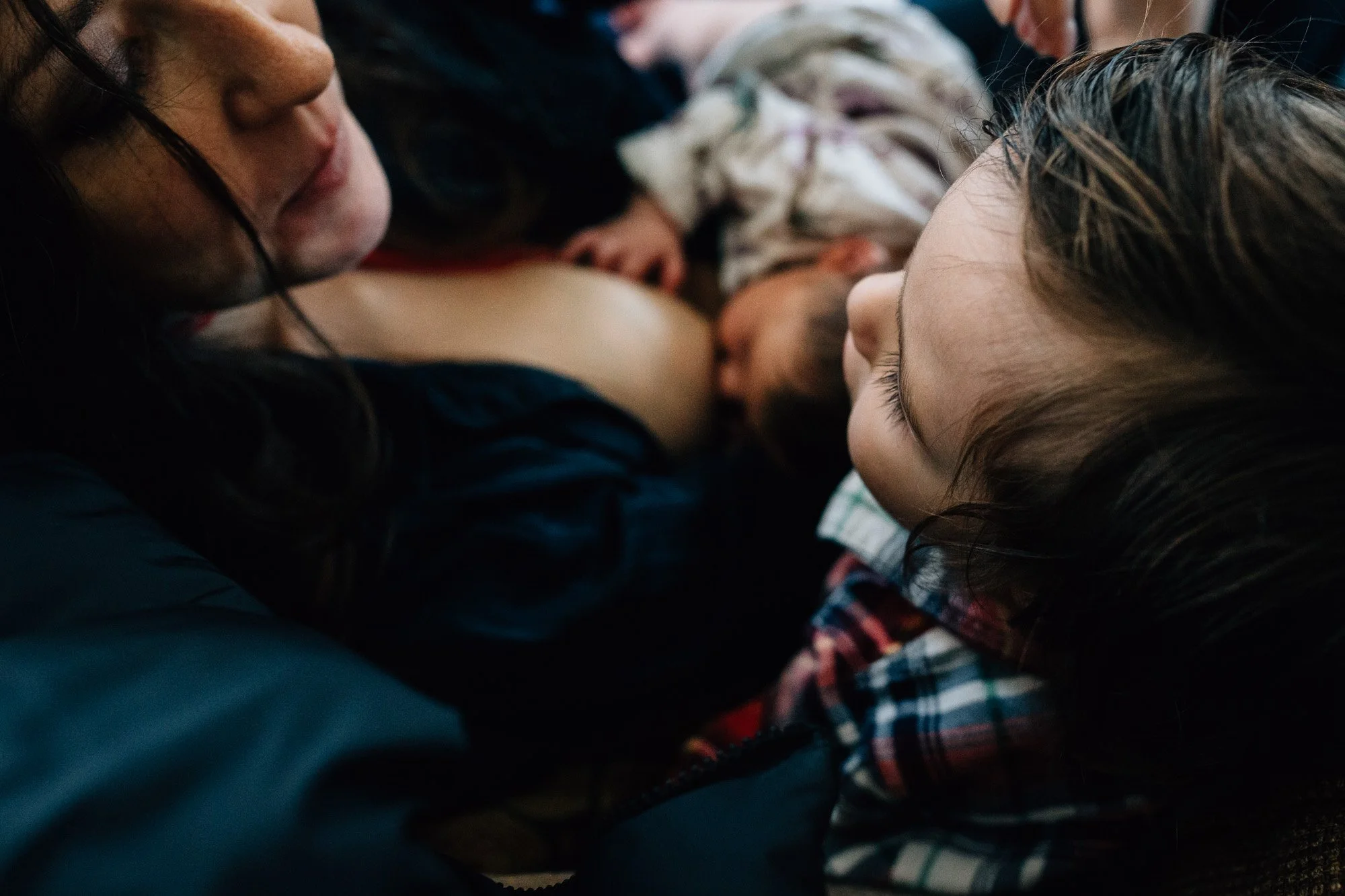 Close-up of two women lying face to face with a baby between them, likely breastfeeding or bonding, in an intimate and tender moment.