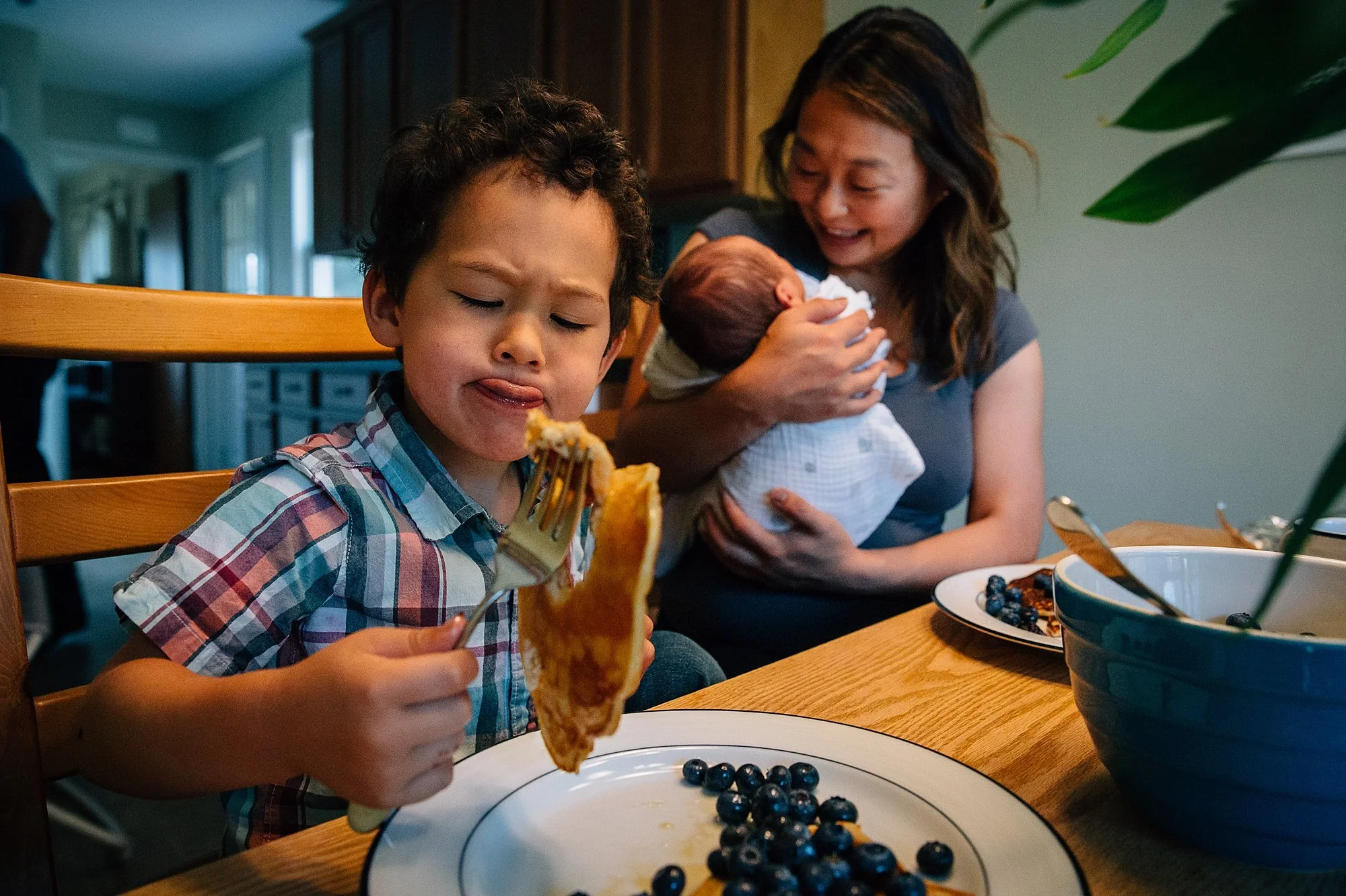 A young boy with curly hair and a plaid shirt making a funny face while eating pancakes at a family meal, with a woman holding a newborn baby in the background.