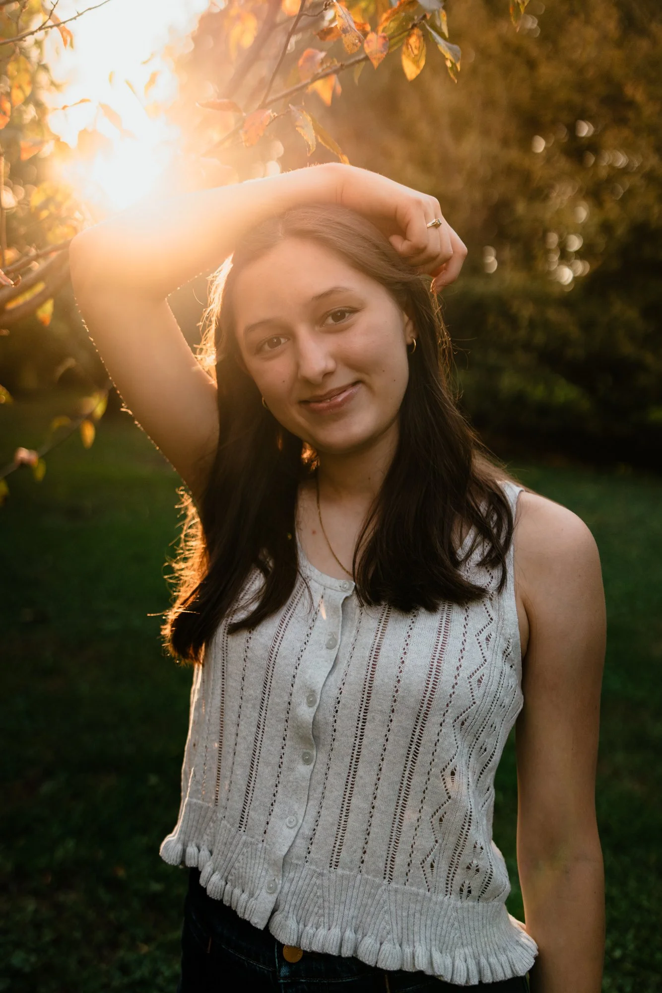 Young woman with brown hair smiling outdoors at sunset, standing near trees with leaves, wearing a sleeveless knit top.