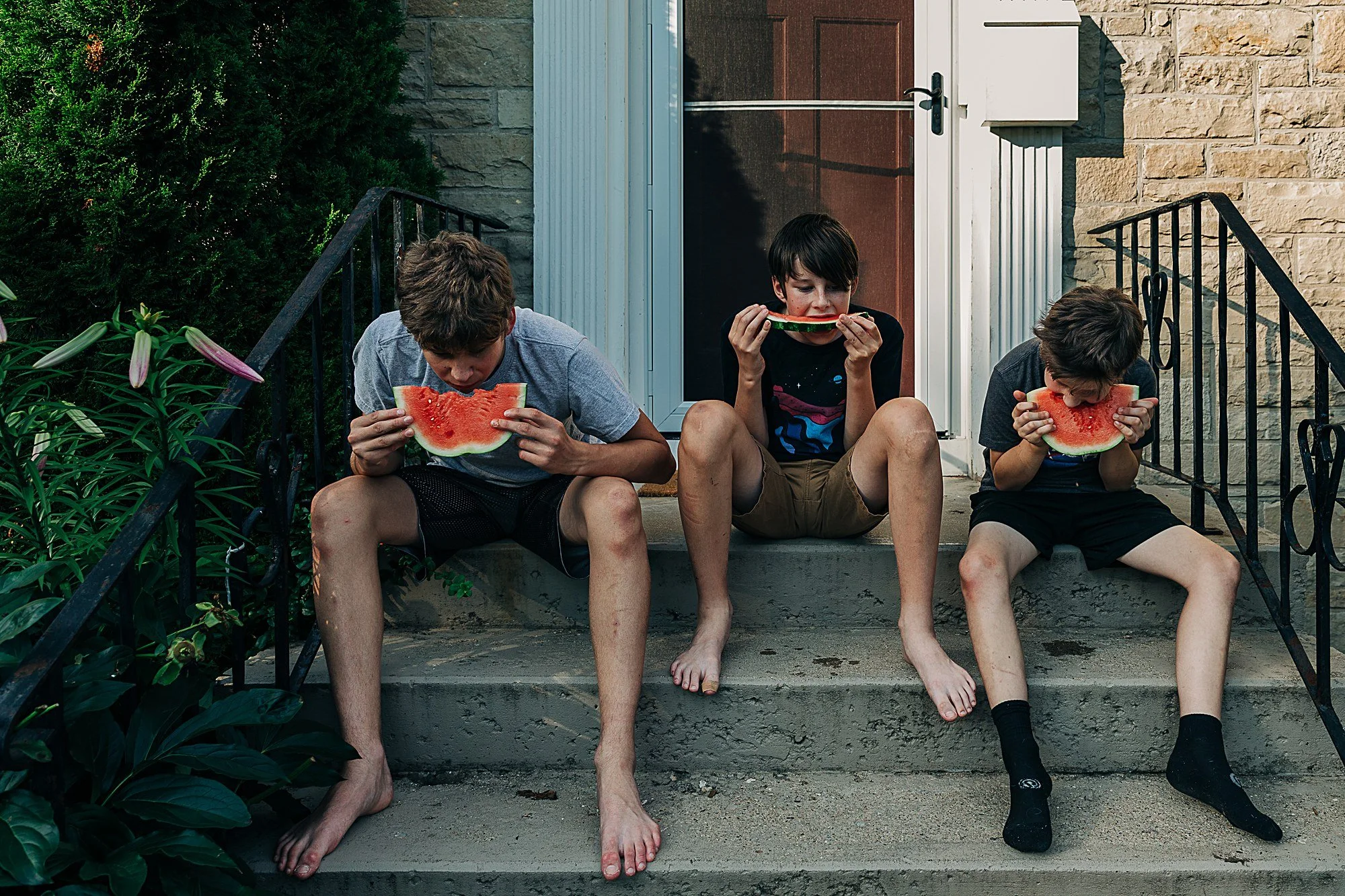 Three boys sitting on the steps outside a house, eating slices of watermelon.