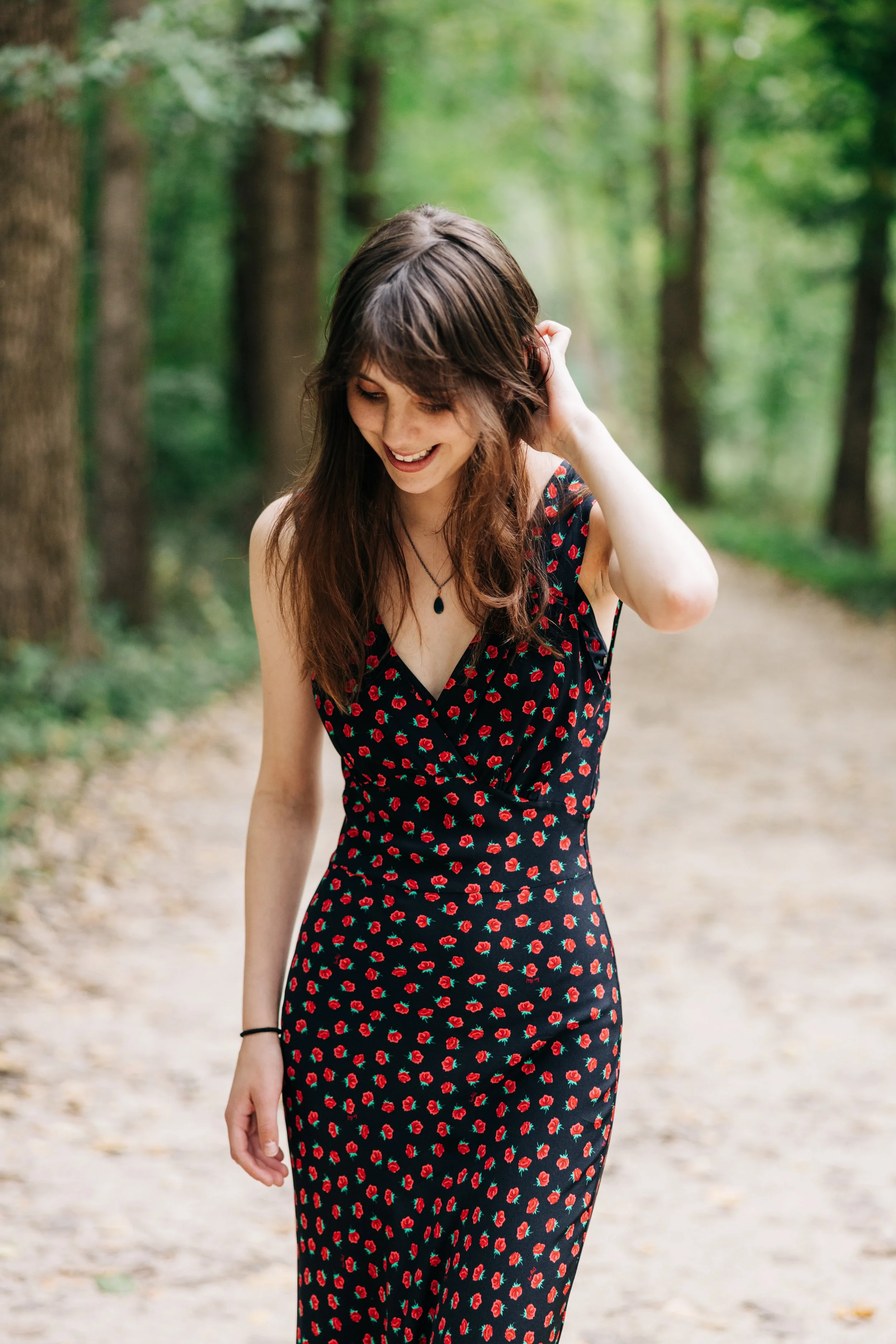 A young woman with long brown hair, wearing a black dress with red floral patterns, walking on a forest path and smiling.