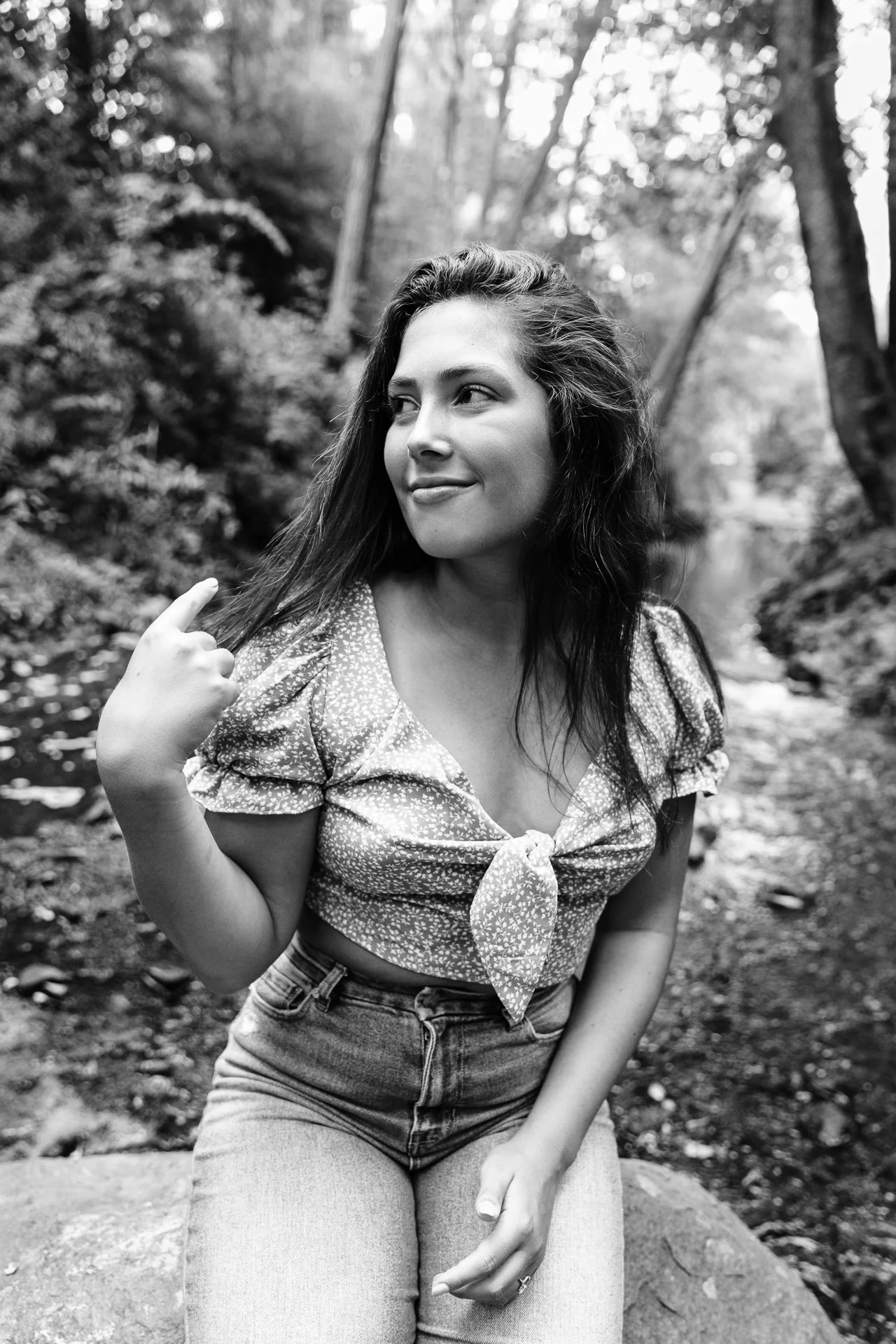 A woman with long hair, wearing a floral-patterned top tied at the front and high-waisted jeans, is sitting outdoors near a small body of water surrounded by trees, looking to the side with a slight smile.