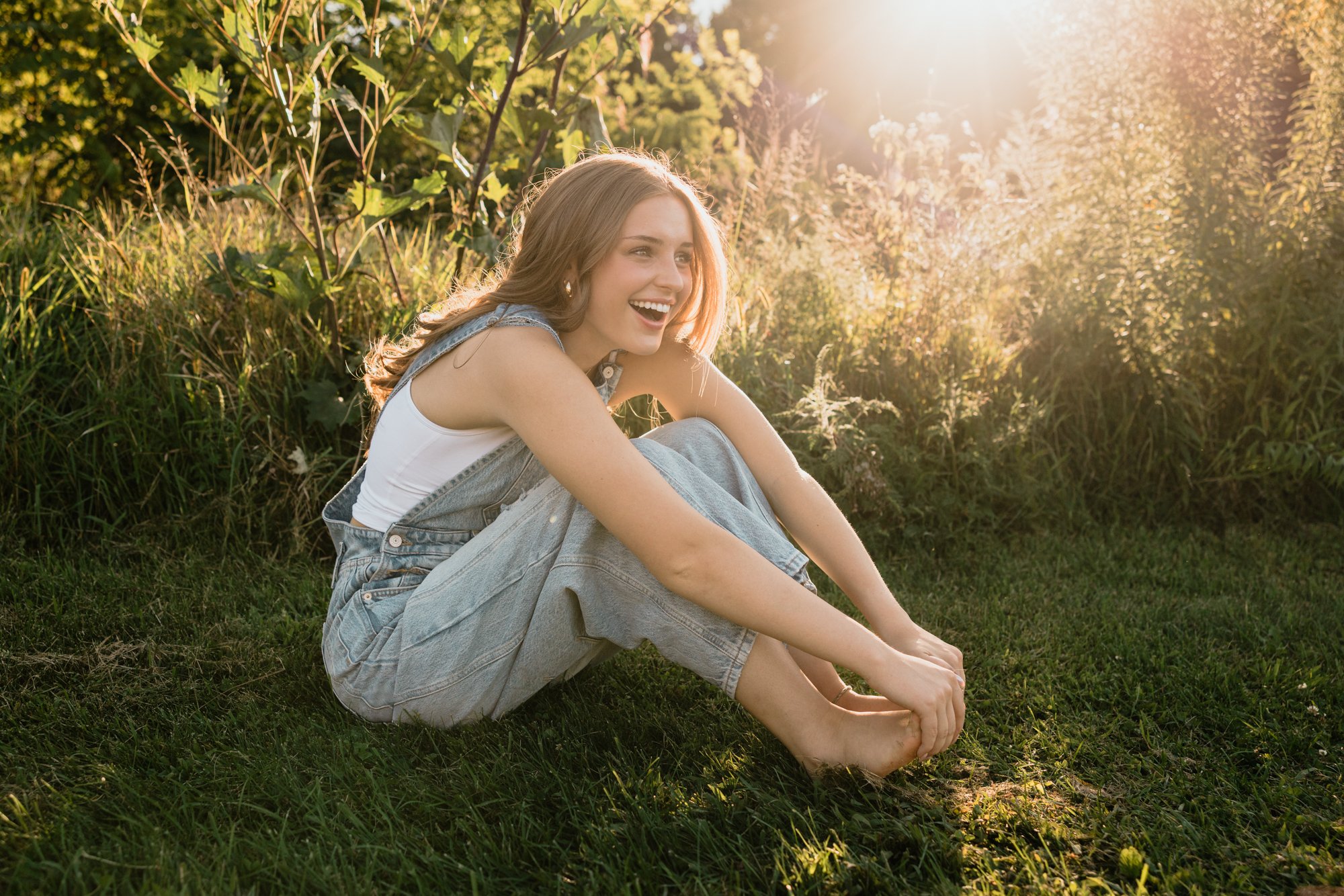 A young woman sitting on grass in a park during sunset, smiling and laughing with her knees drawn up, wearing a white tank top and light denim overalls.