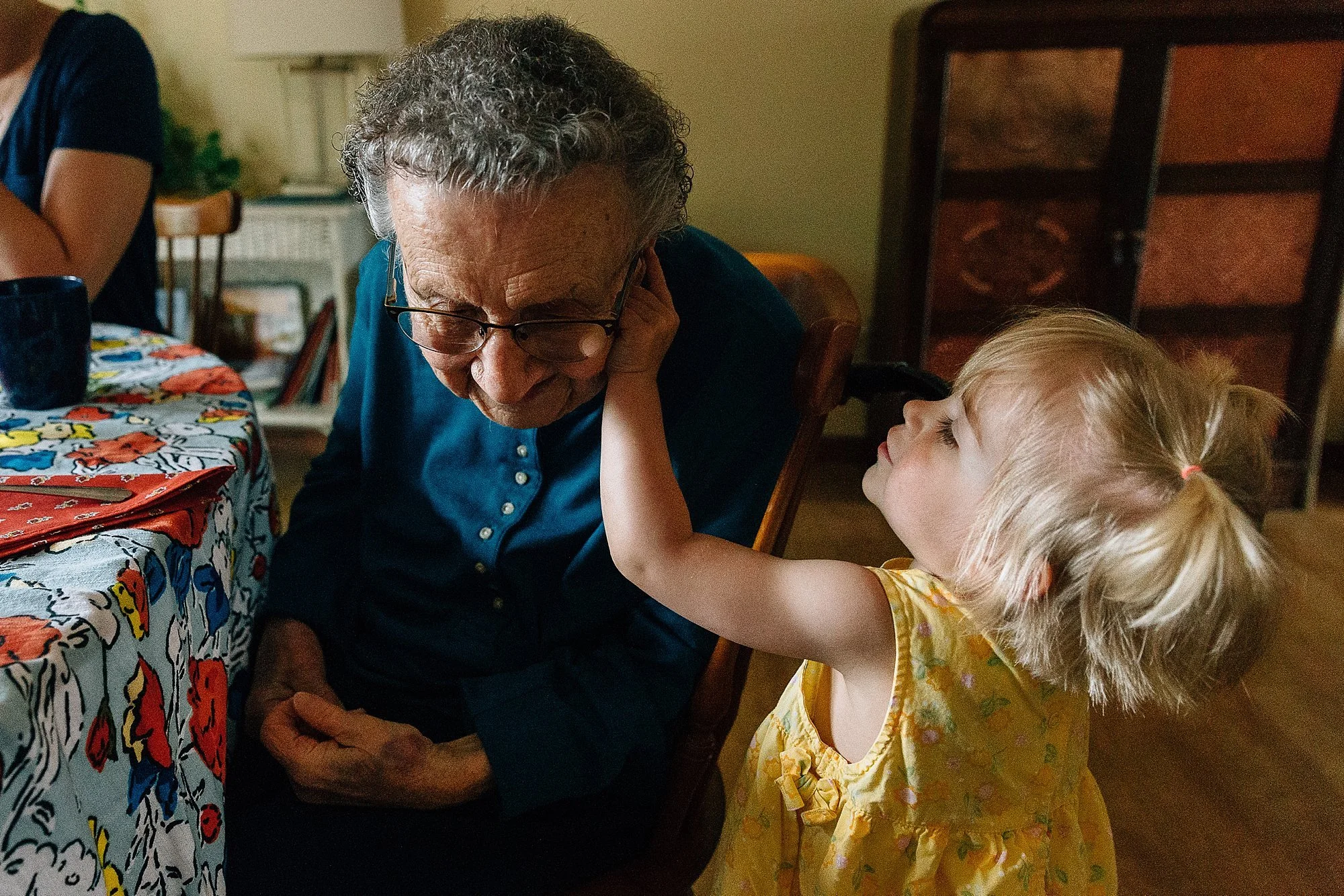 A young girl with blonde hair and two pigtails touching an elderly woman's face at a dining table.