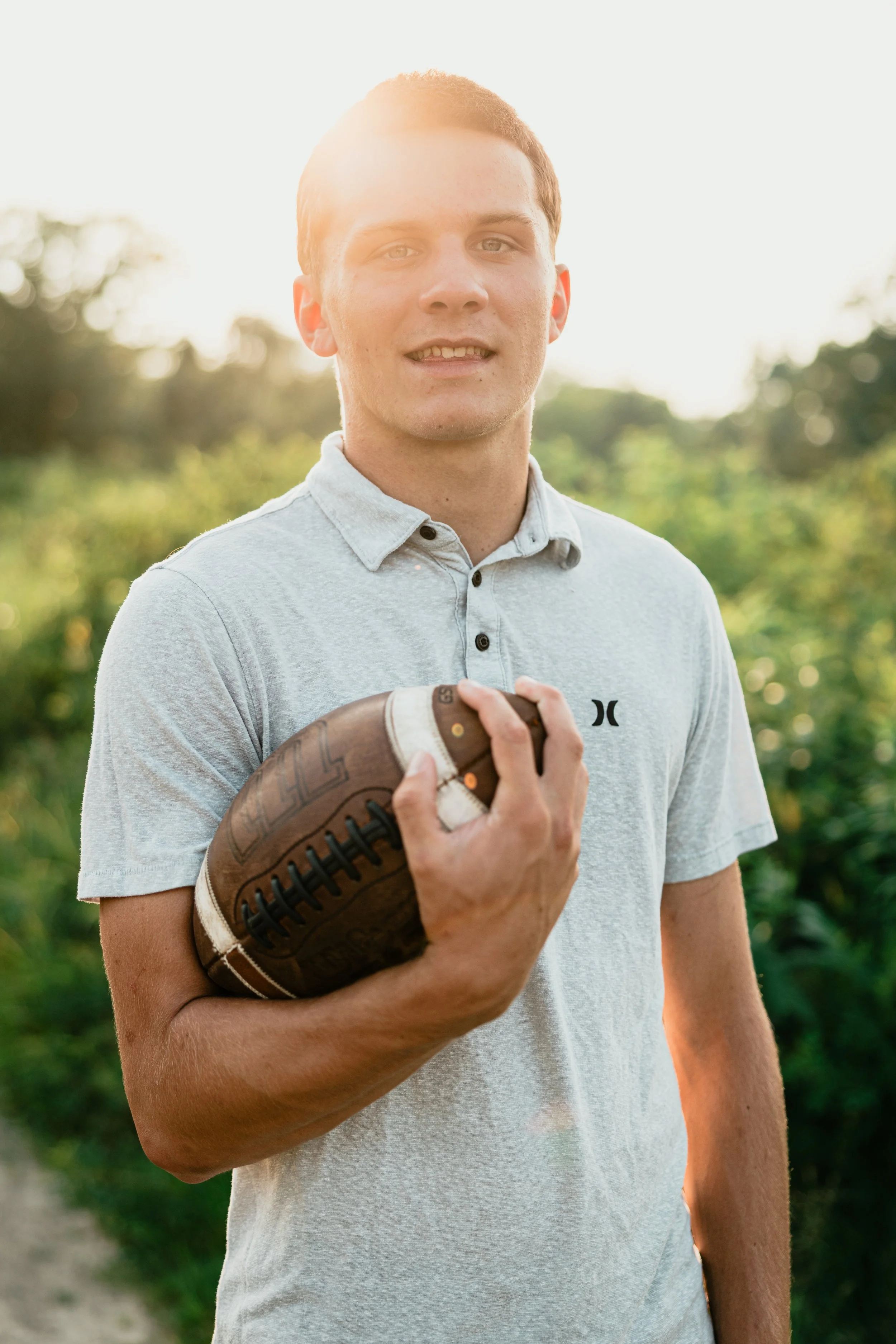 A young man outdoors holding a football, wearing a light gray polo shirt, with sunlight and greenery in the background.