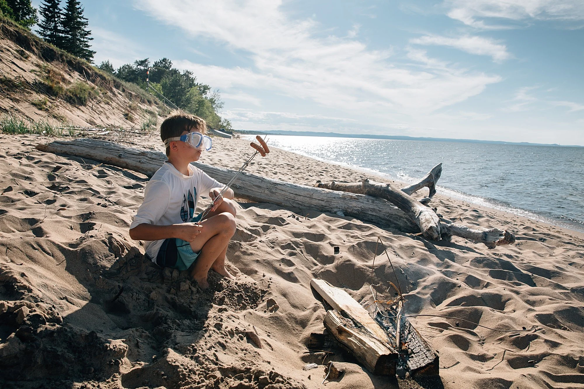 A young boy sitting on a sandy beach near the water, wearing goggles and holding a stick, with driftwood around him and trees in the background.