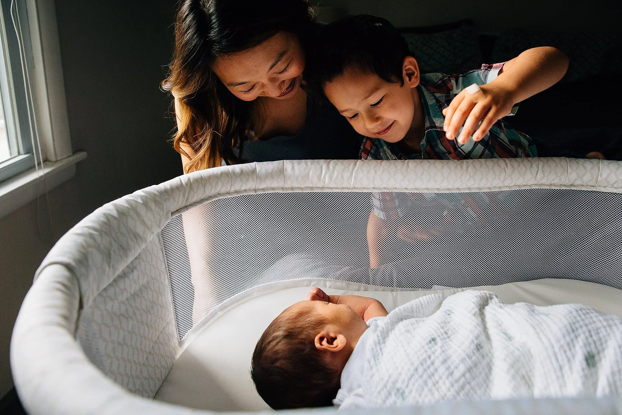 A woman and a young boy look into a crib at a sleeping baby wrapped in a white blanket.