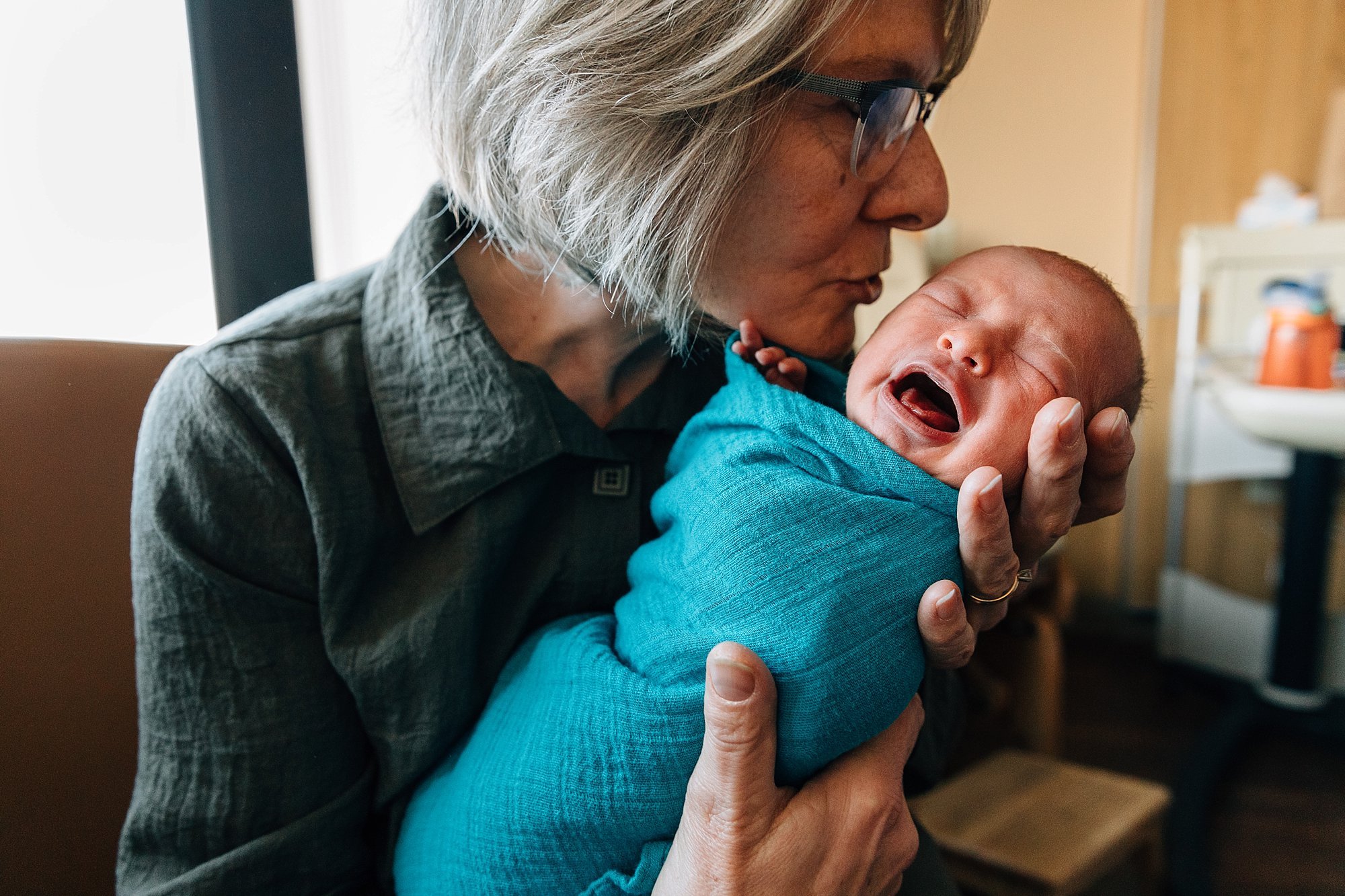 An older woman holding and kissing a crying baby wrapped in a blue blanket indoors.