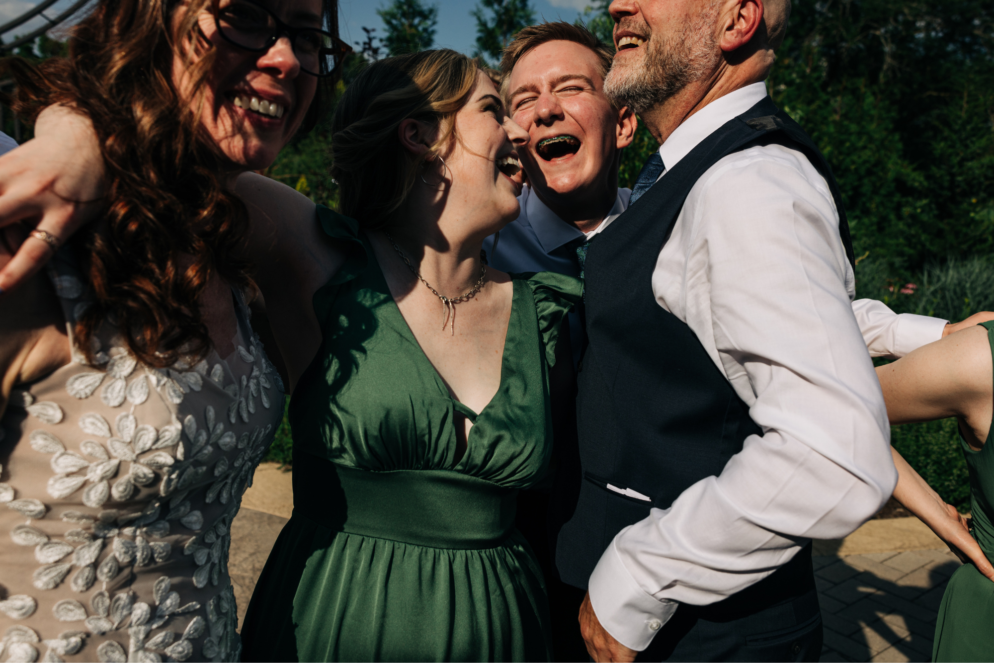 Group of five people smiling and laughing close together outdoors during a sunny day.