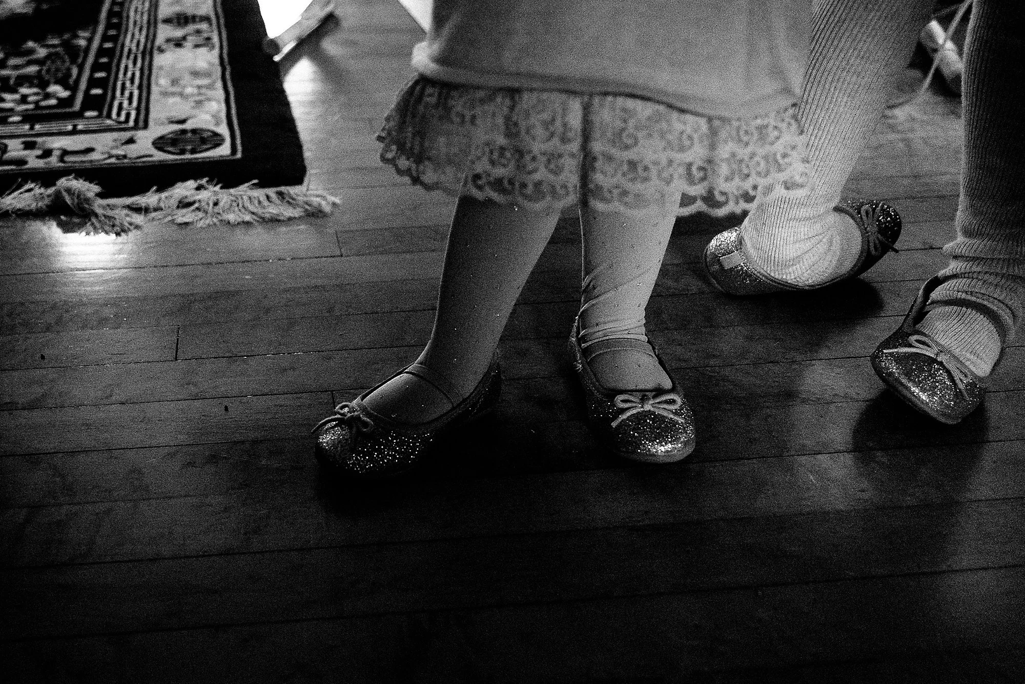Close-up of children's legs and feet wearing sparkly shoes, tights, and dresses on a wooden floor, with a decorative rug nearby.