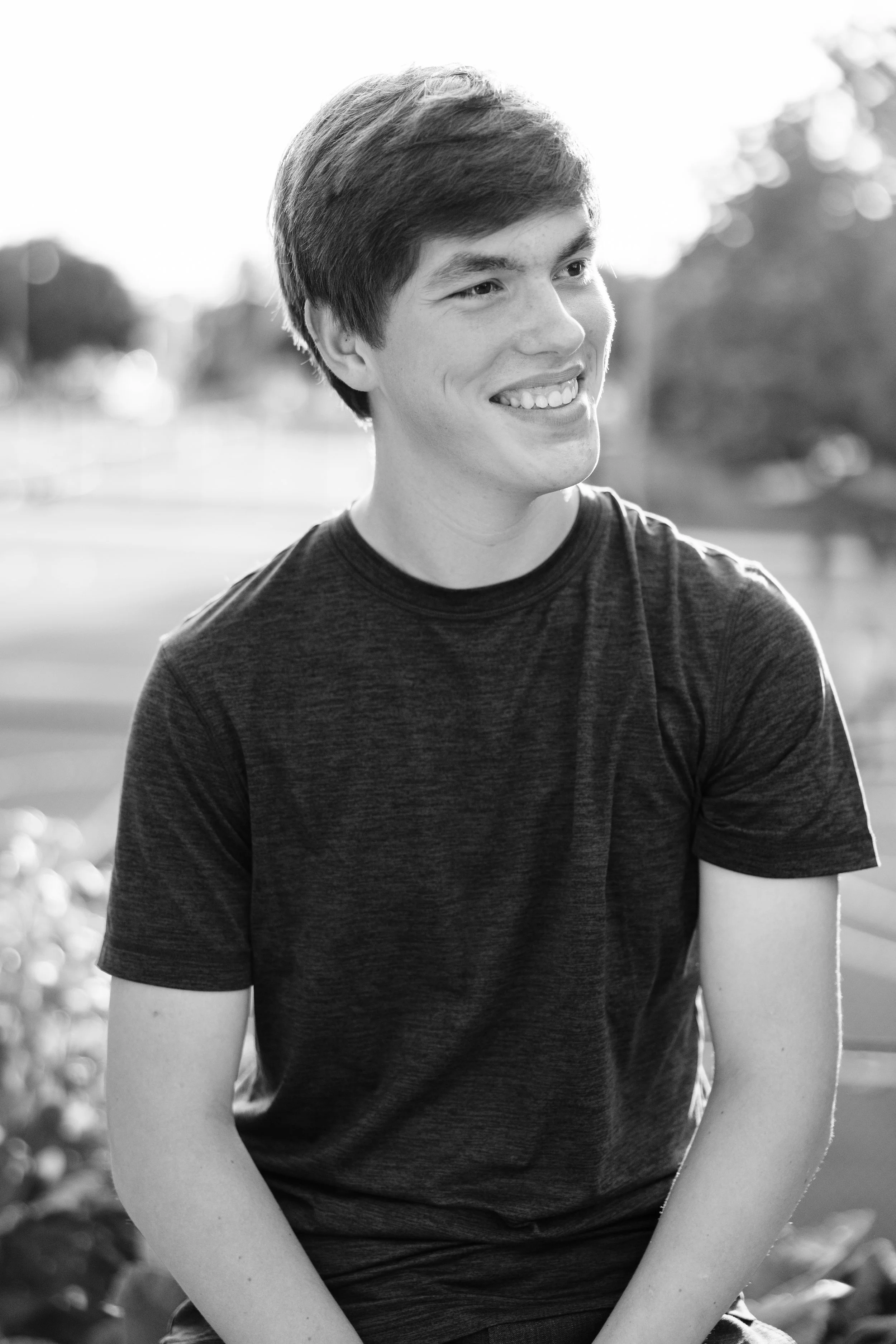 A young man smiling and sitting outdoors, wearing a dark t-shirt, with a background of trees and a park.
