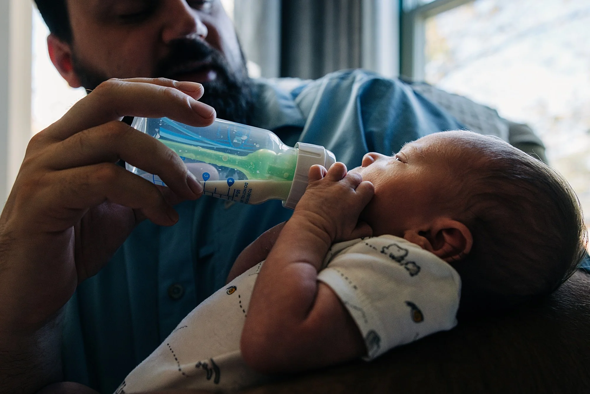 An adult man feeding a baby with a bottle of milk inside a room with sunlight coming through a window.