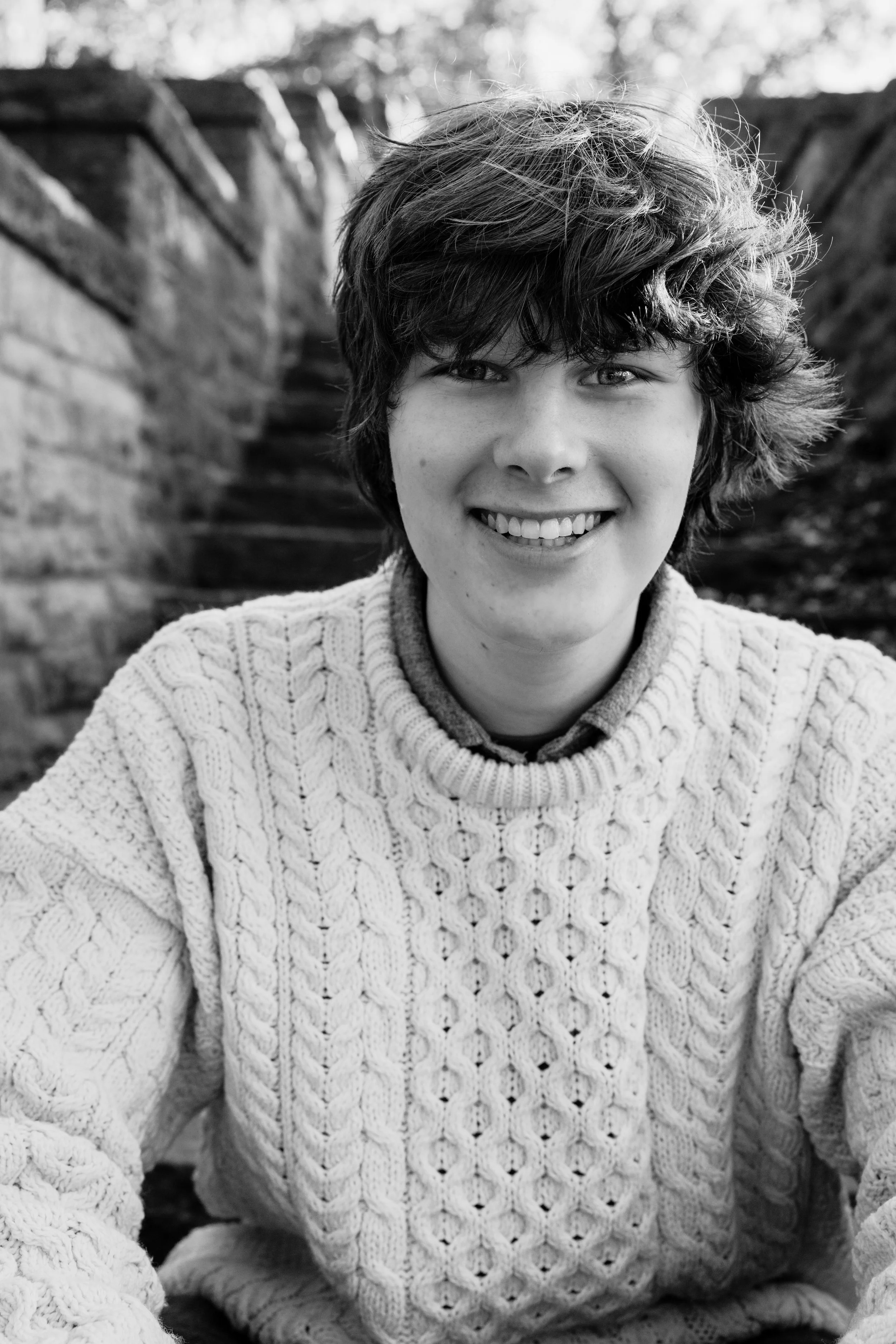 Smiling young person with wavy hair wearing a cable-knit sweater, outdoors with stone steps and a stone wall in the background