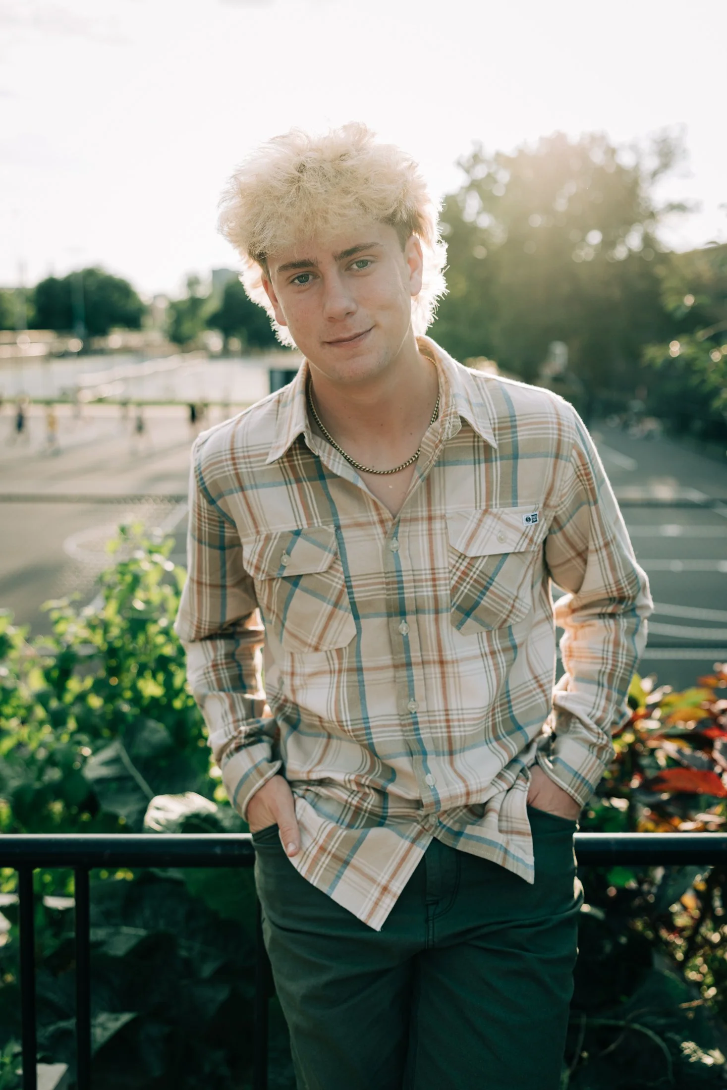 A young man with blond, curly hair stands outdoors in the late afternoon with trees and a parking lot in the background, wearing a plaid shirt and black pants.