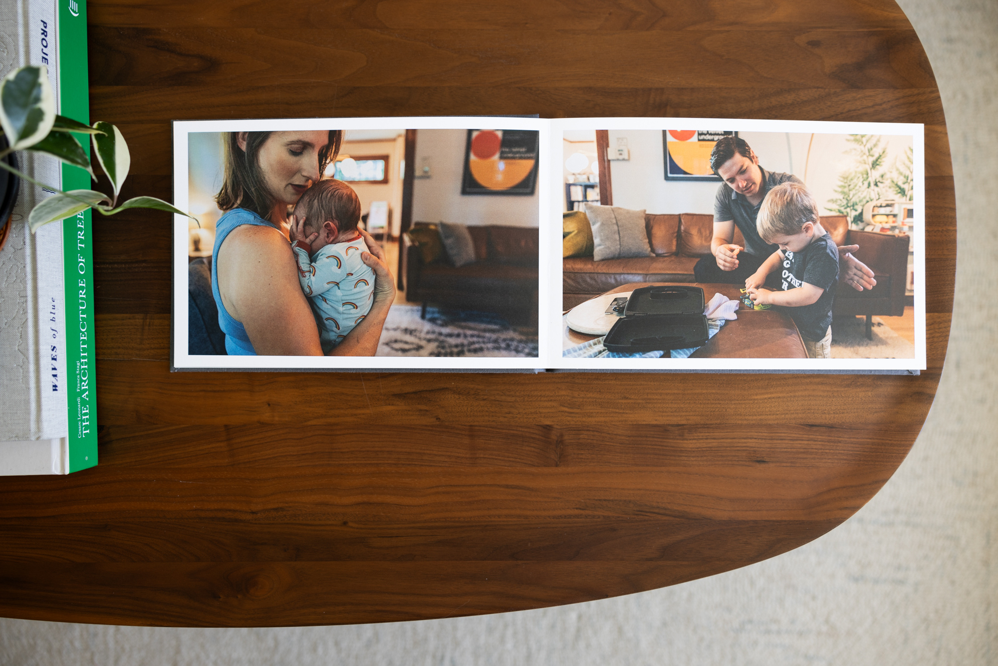 Open photo album displaying two photos of family moments: one of a woman holding a newborn baby, and the other of a man and young boy playing with toys on a couch in a living room.
