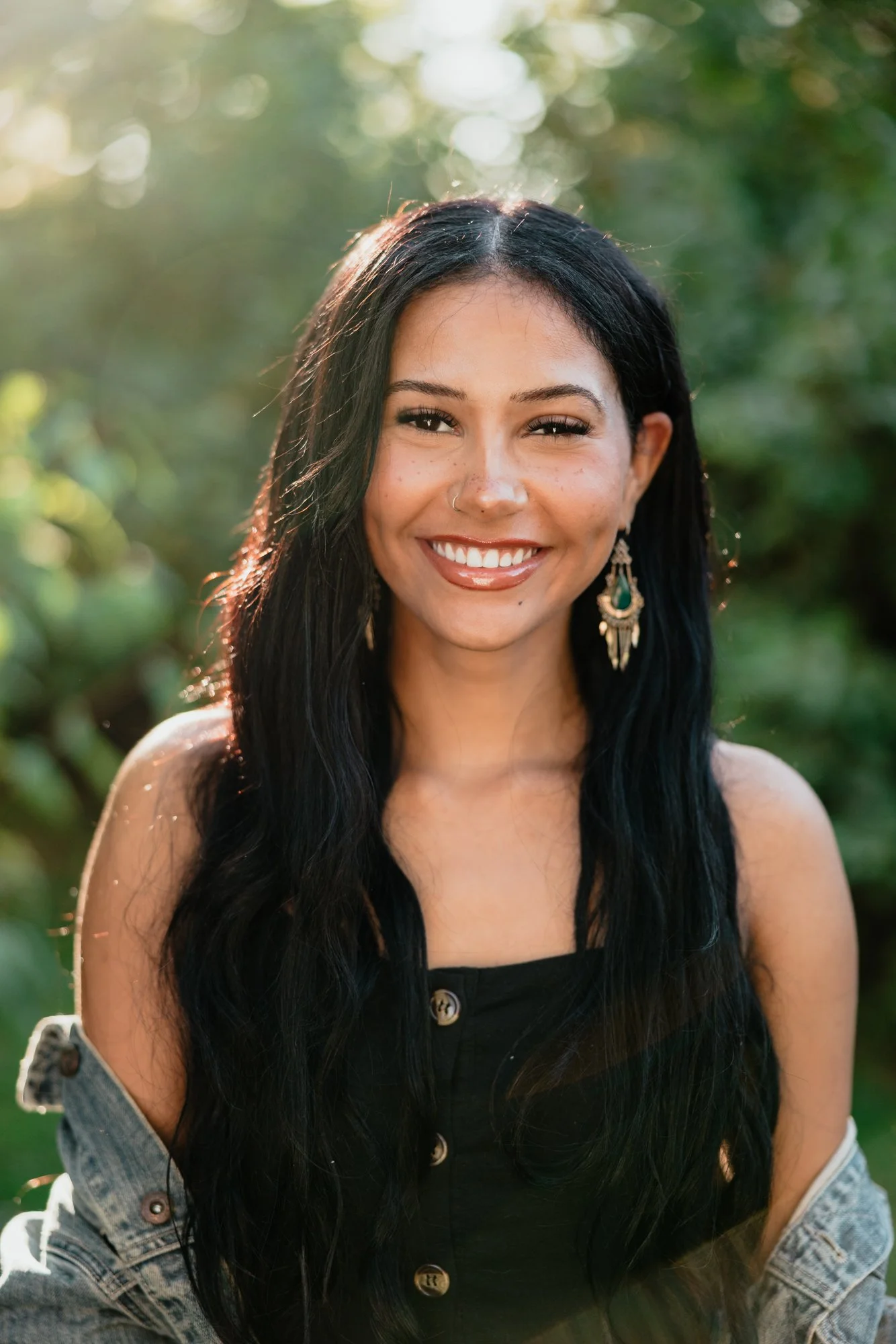 A woman with long black hair, wearing earrings and a black top, smiling outdoors with sunlight and greenery in the background.