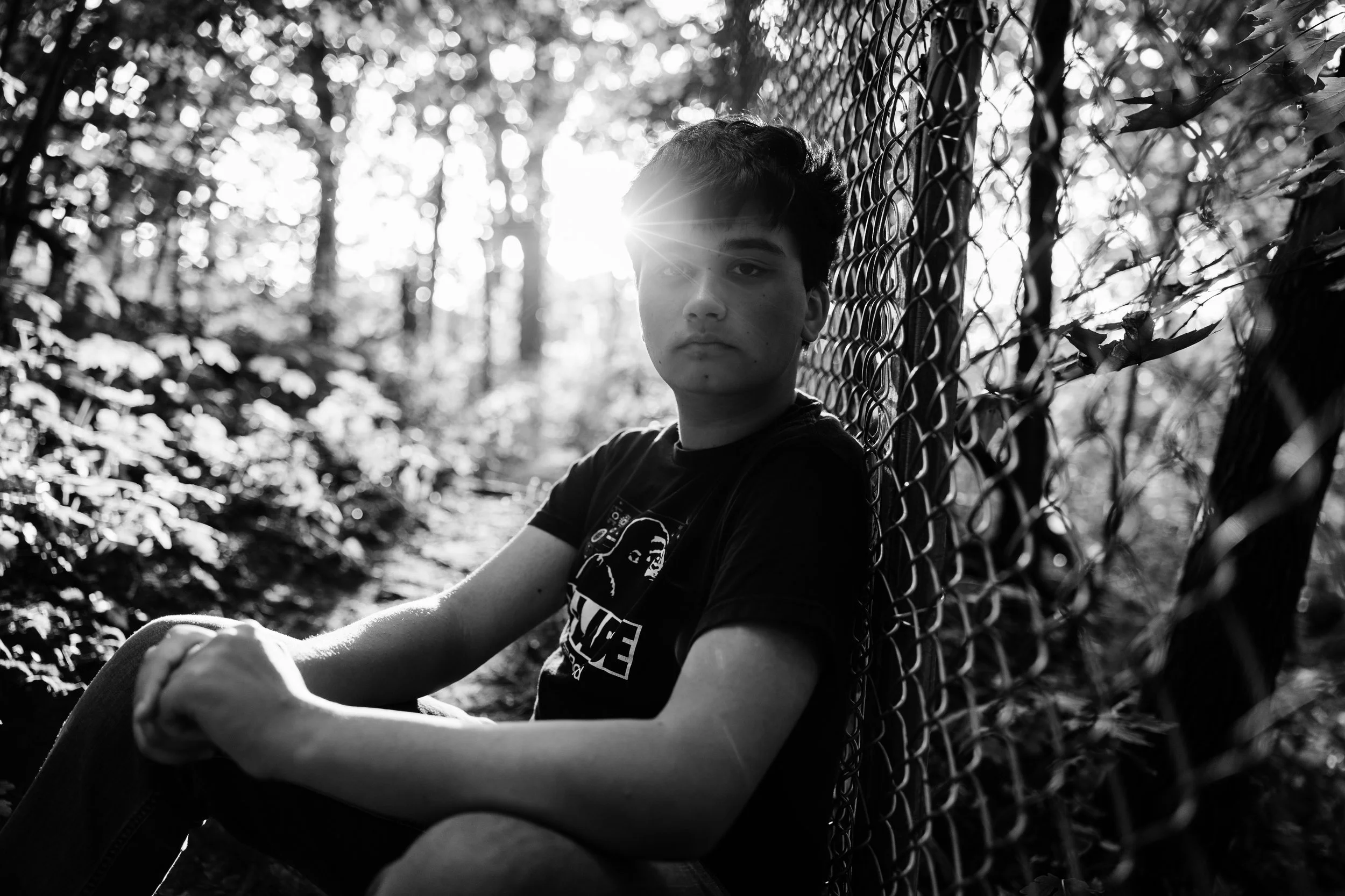 A young person sitting against a chain-link fence in a forest, with sunlight shining through the trees behind them, captured in black and white.
