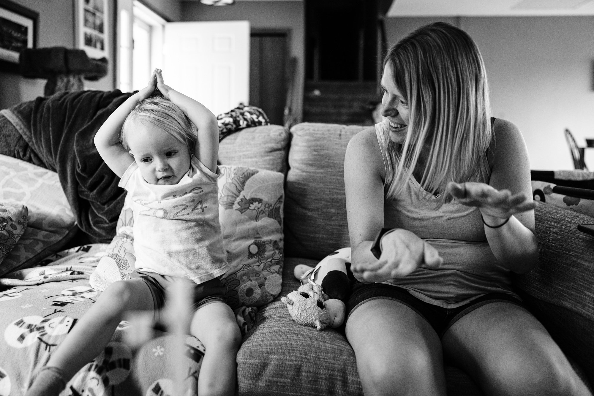 A young girl and an adult woman sitting on a couch having a conversation in a cozy living room, with the girl making a playful gesture with her arms above her head.